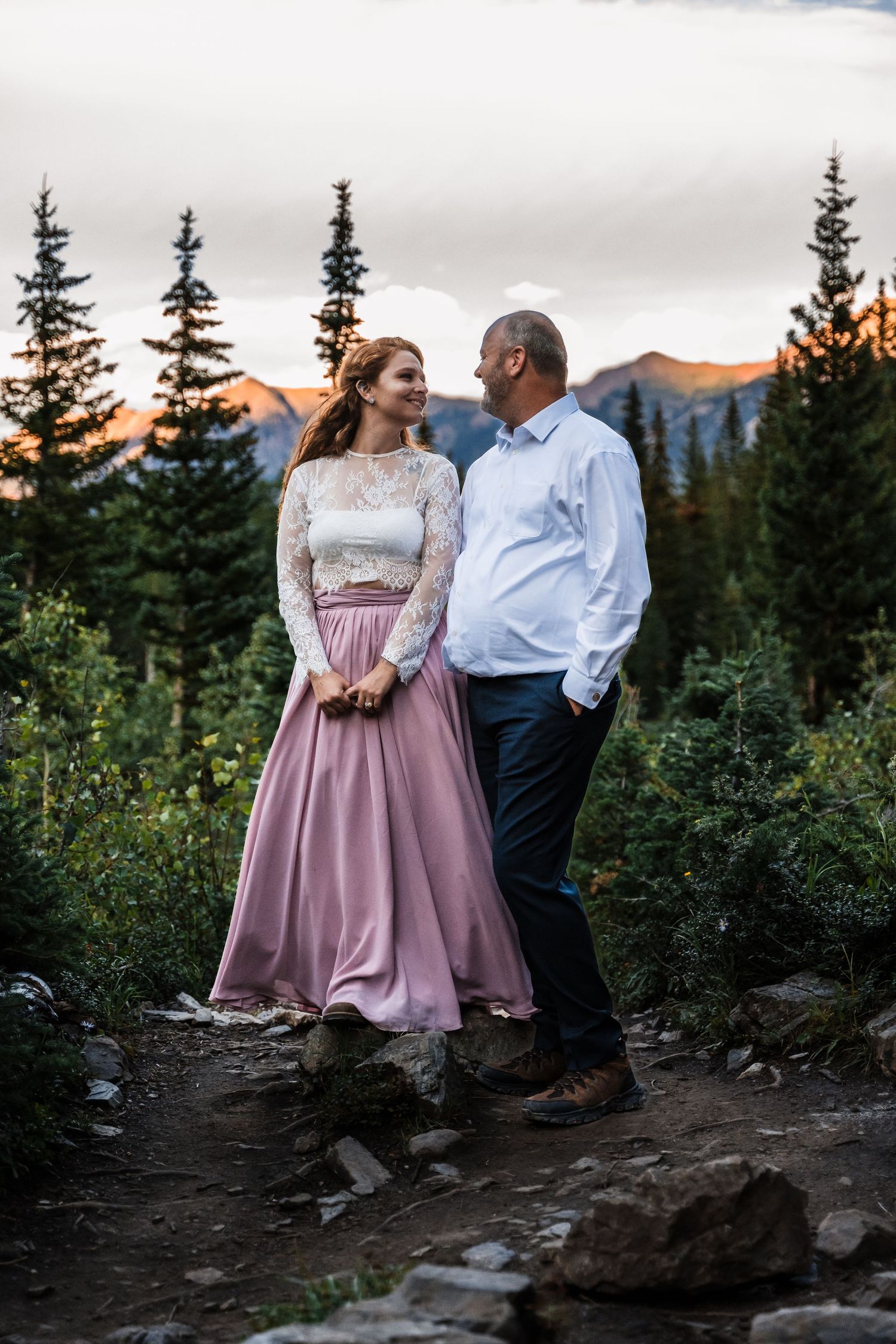 Elegant couple in formal attire standing on mountain trail at dusk.