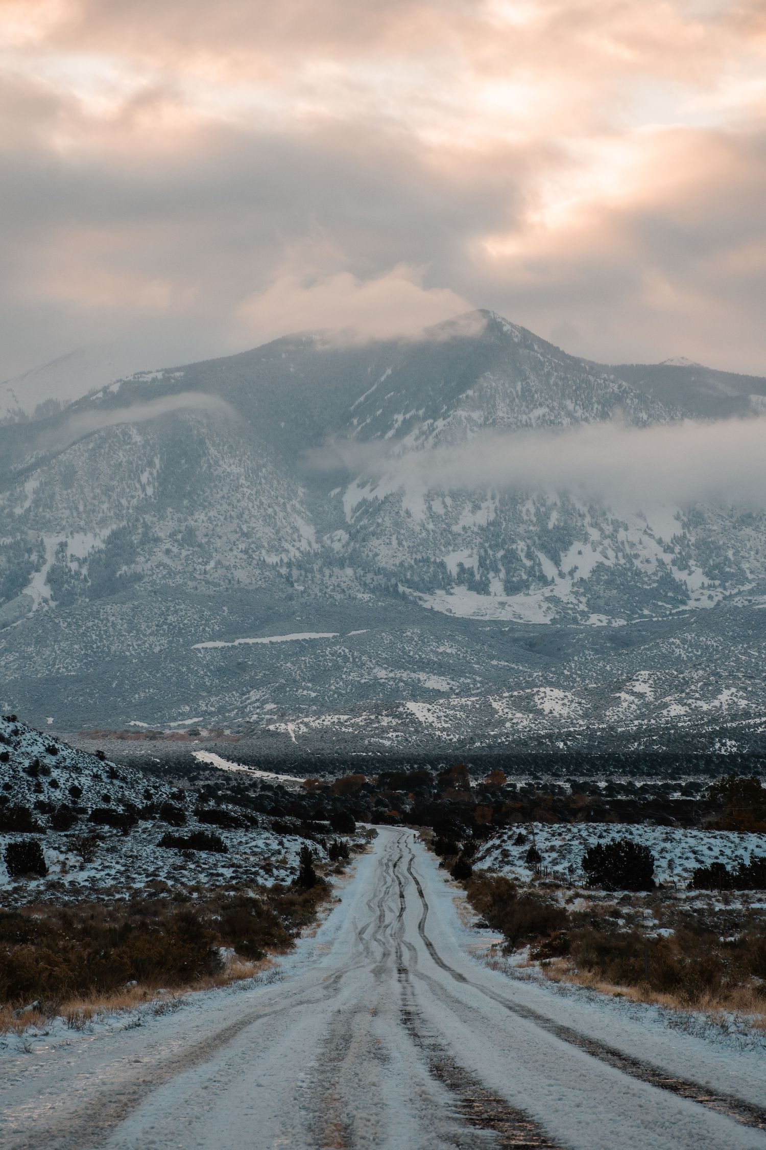 A snowy dirt road leads toward a majestic mountain peak shrouded in moody winter clouds.