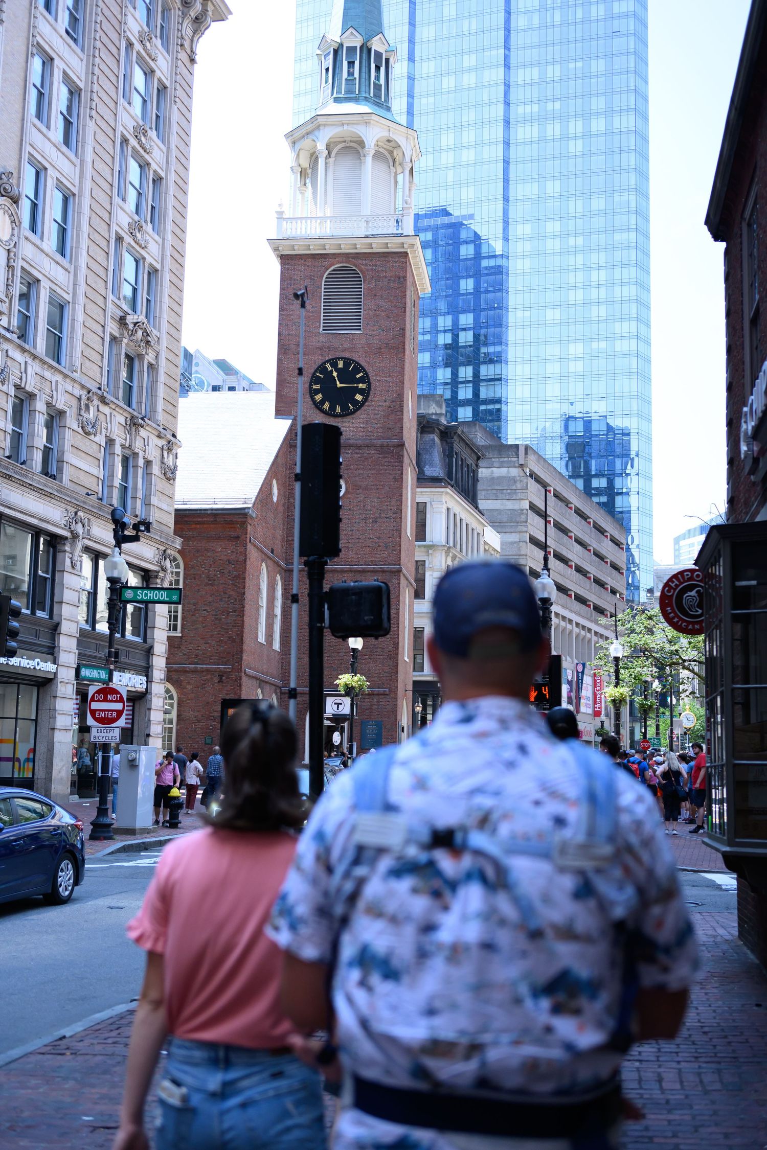 Two people walk past the historic Old South Meeting House clock tower in downtown Boston with modern skyscrapers in background.
