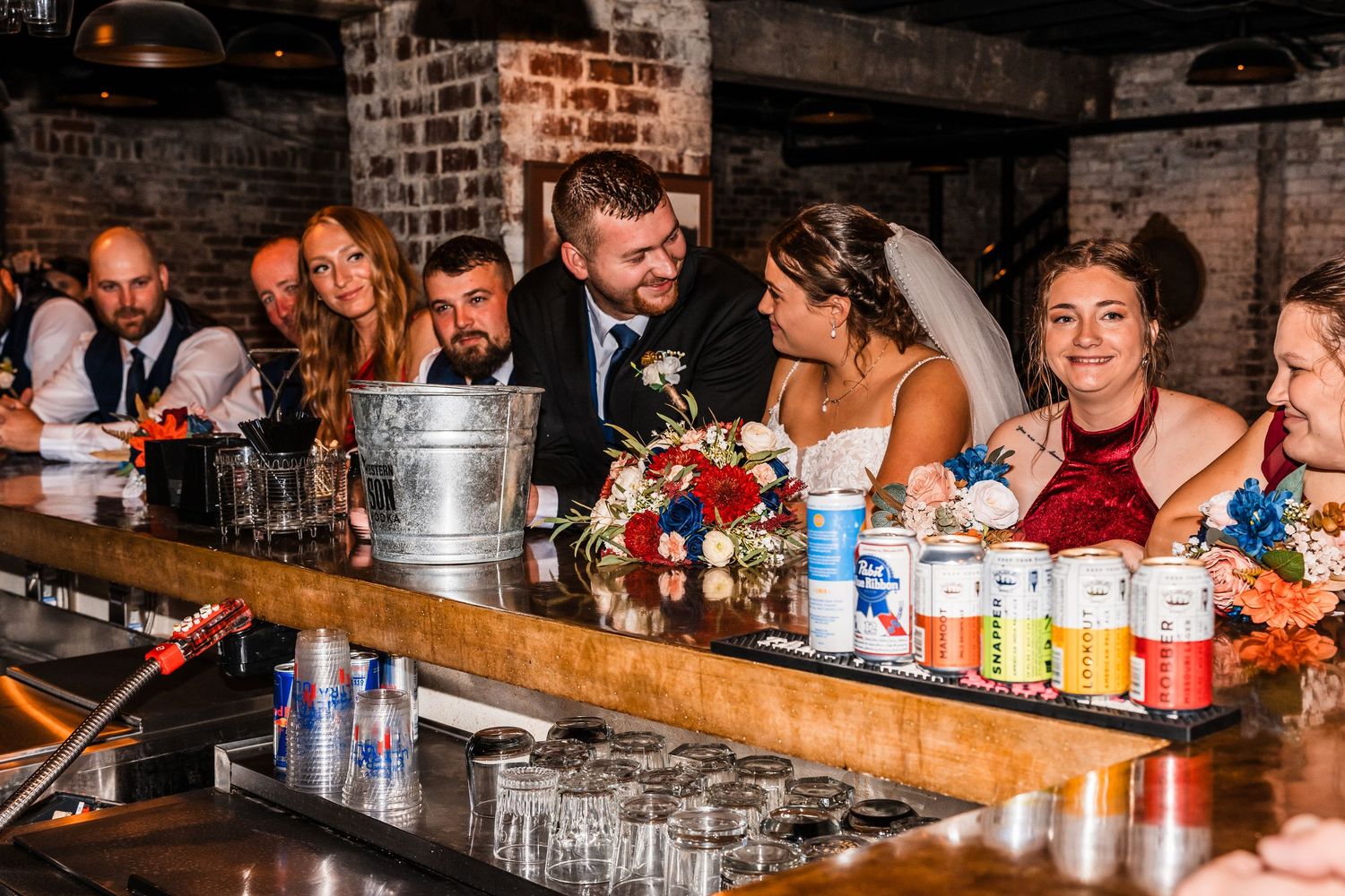 Wedding guests enjoying drinks and conversation at a rustic brick-walled bar during reception celebration.