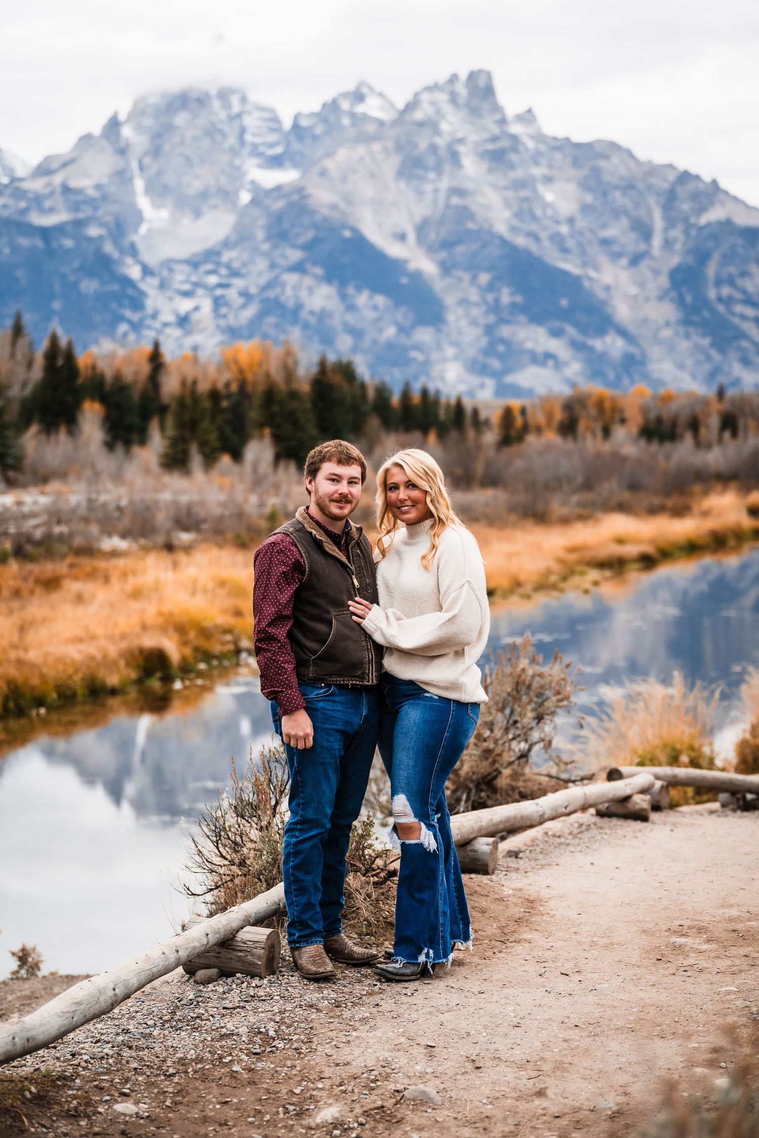 A couple stands together on a trail overlooking a river with the Grand Teton mountains rising majestically in the background.