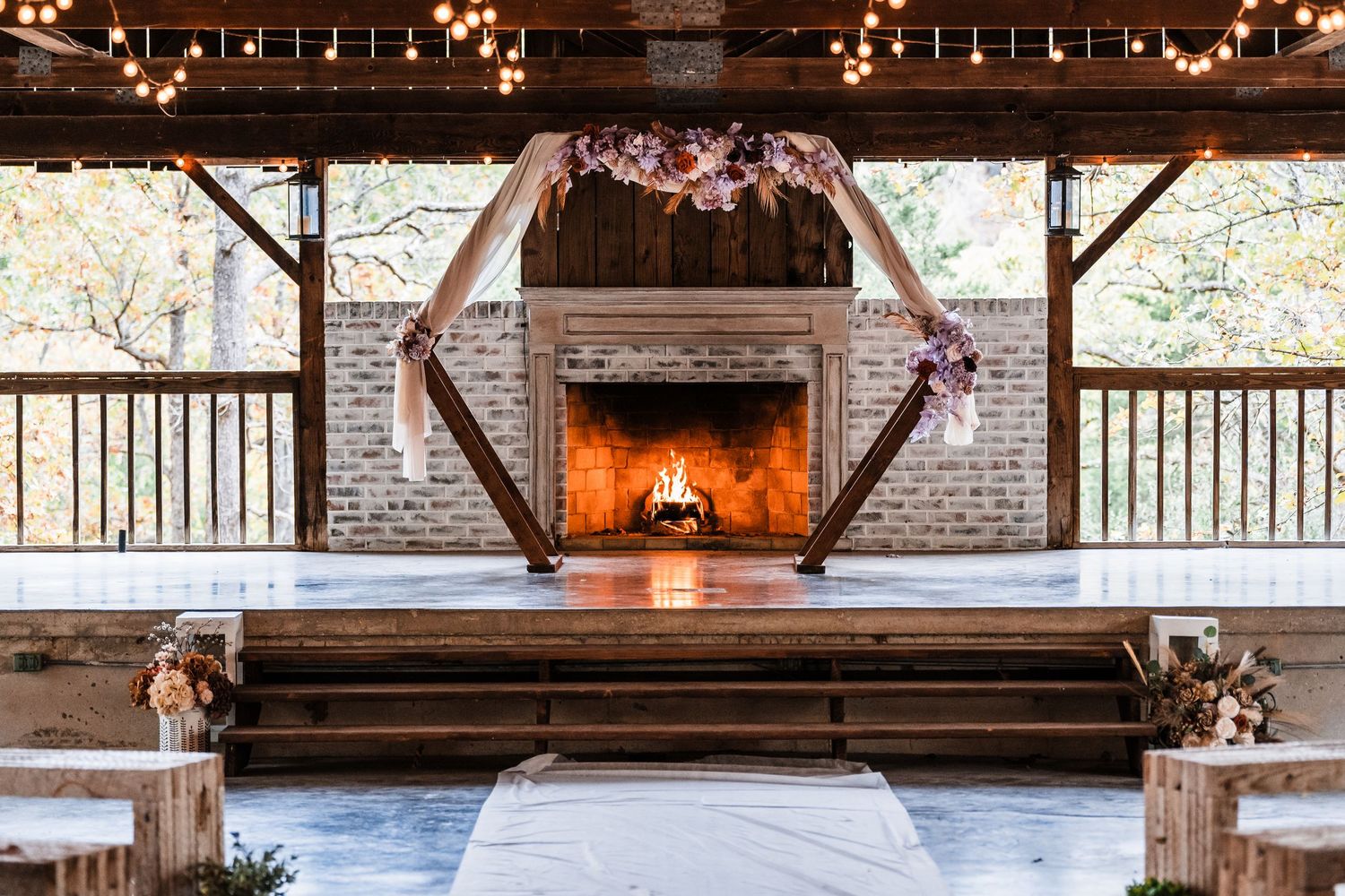 A rustic wedding ceremony setup with hexagonal arch and fireplace decorated with string lights.