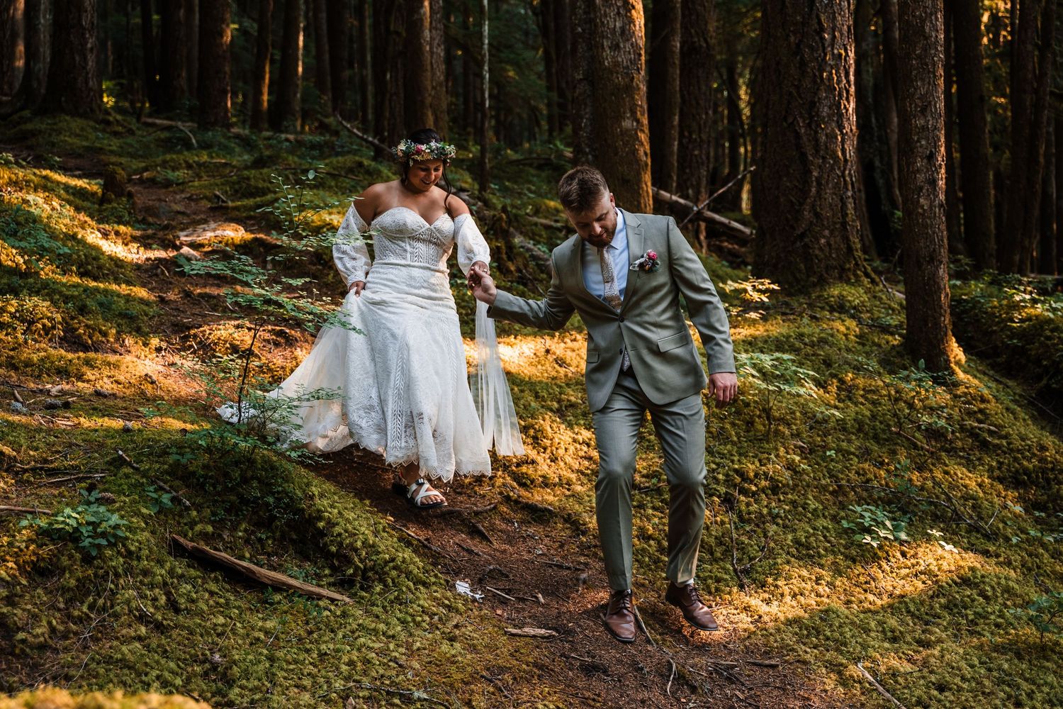 A couple in wedding attire walks hand in hand through a sunlit forest path during golden hour.