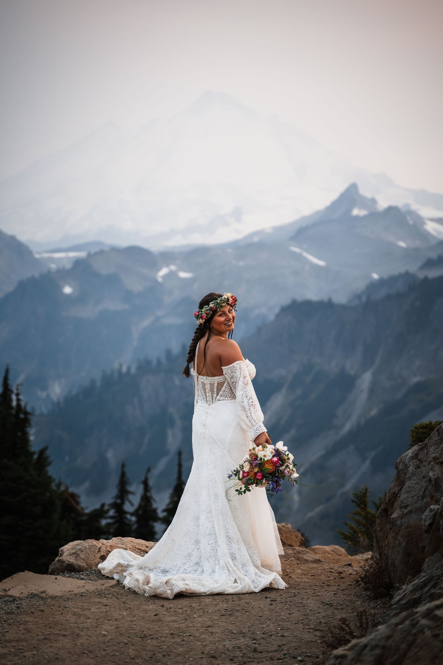 A bride in a white lace wedding dress sits on a mountain summit overlooking a dramatic mountain range at sunset.