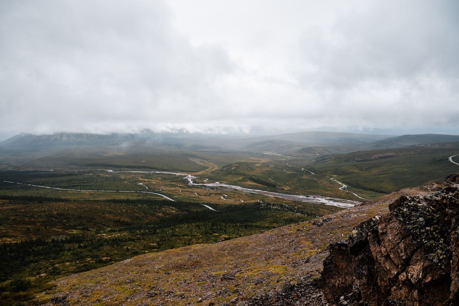 Winding mountain road cuts through rugged terrain with foggy peaks and valleys in the distance under moody skies.