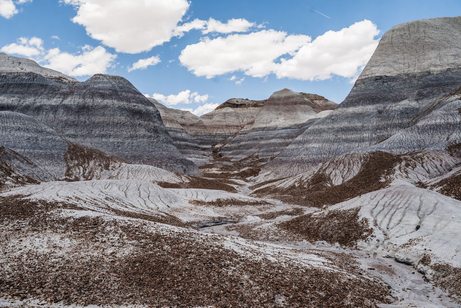 Layered sedimentary rock formations create dramatic striped patterns in a barren desert landscape beneath puffy clouds.