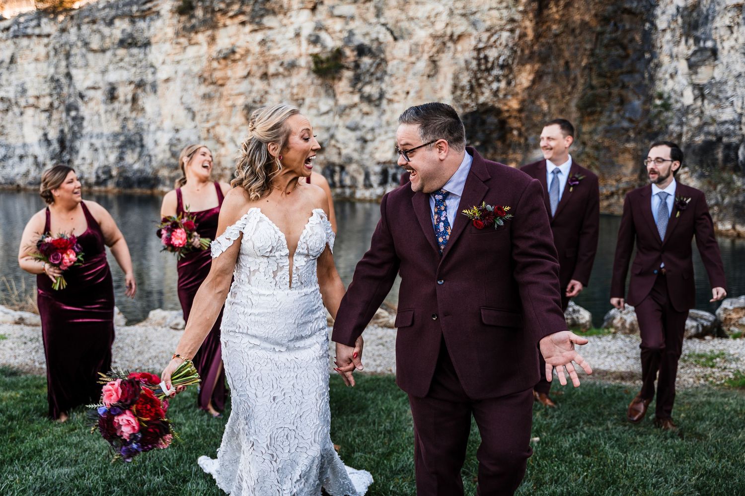 Wedding party in burgundy suits and white dress pose against rustic stone wall during autumn celebration.
