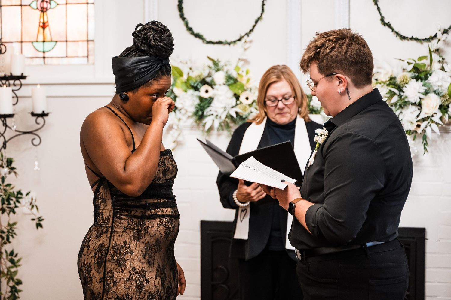 A couple shares an emotional moment during their wedding ceremony with an officiant at a venue decorated with white flowers.