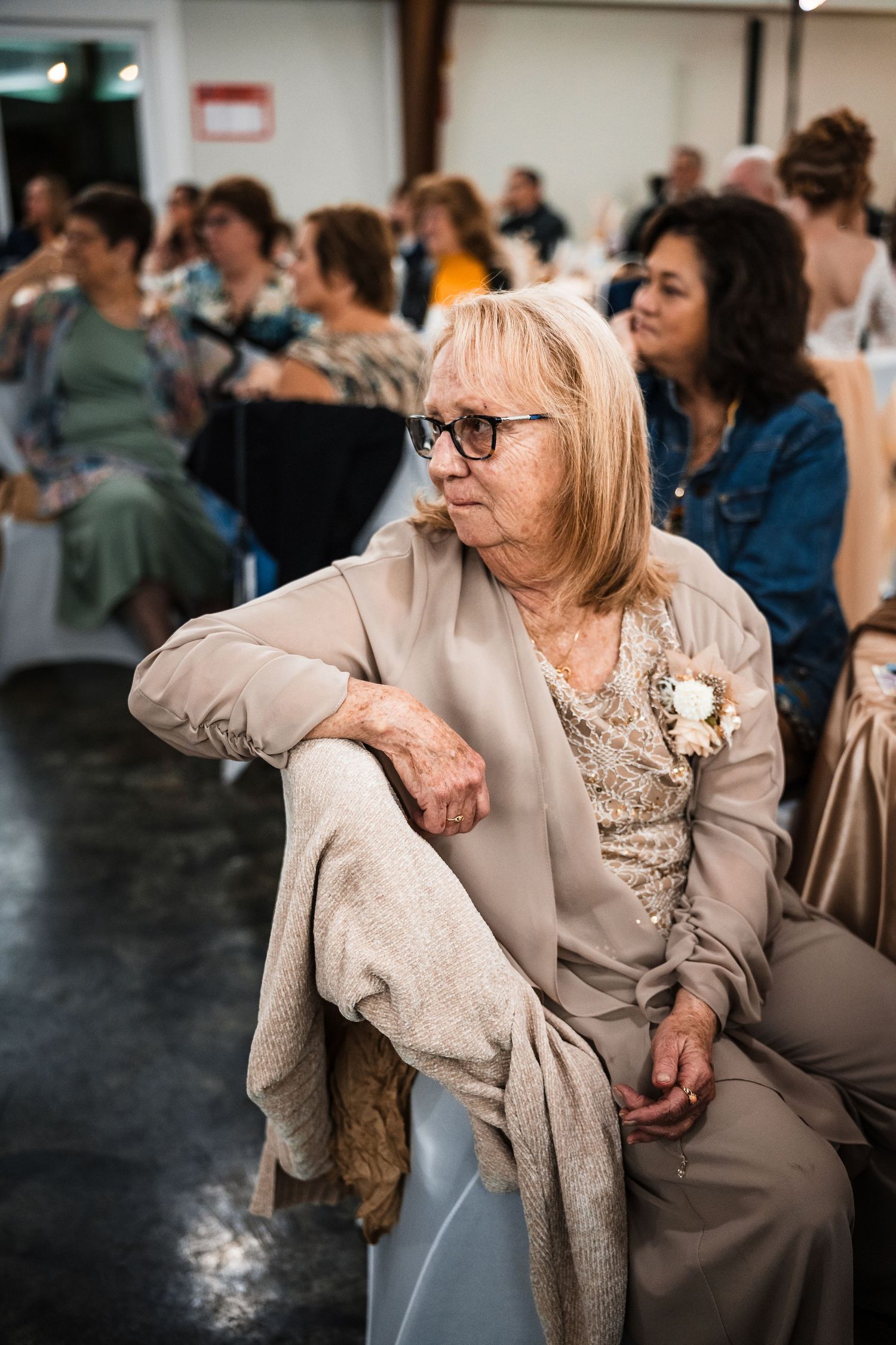 An attendee in glasses and beige attire sits thoughtfully during an indoor event with others in the background.