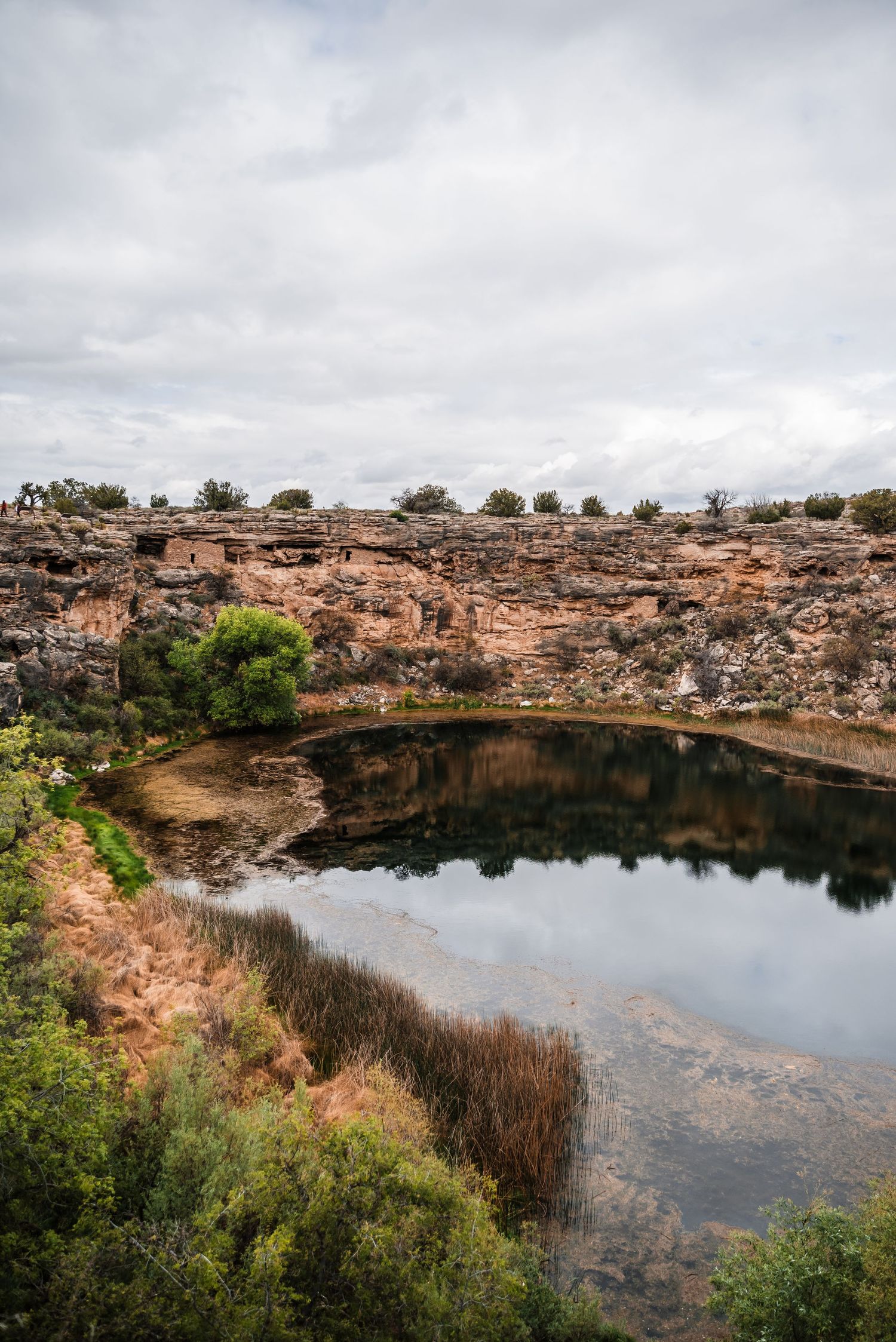 A natural sinkhole or cenote with still water reflects rocky cliffs and vegetation under cloudy skies in a desert landscape.