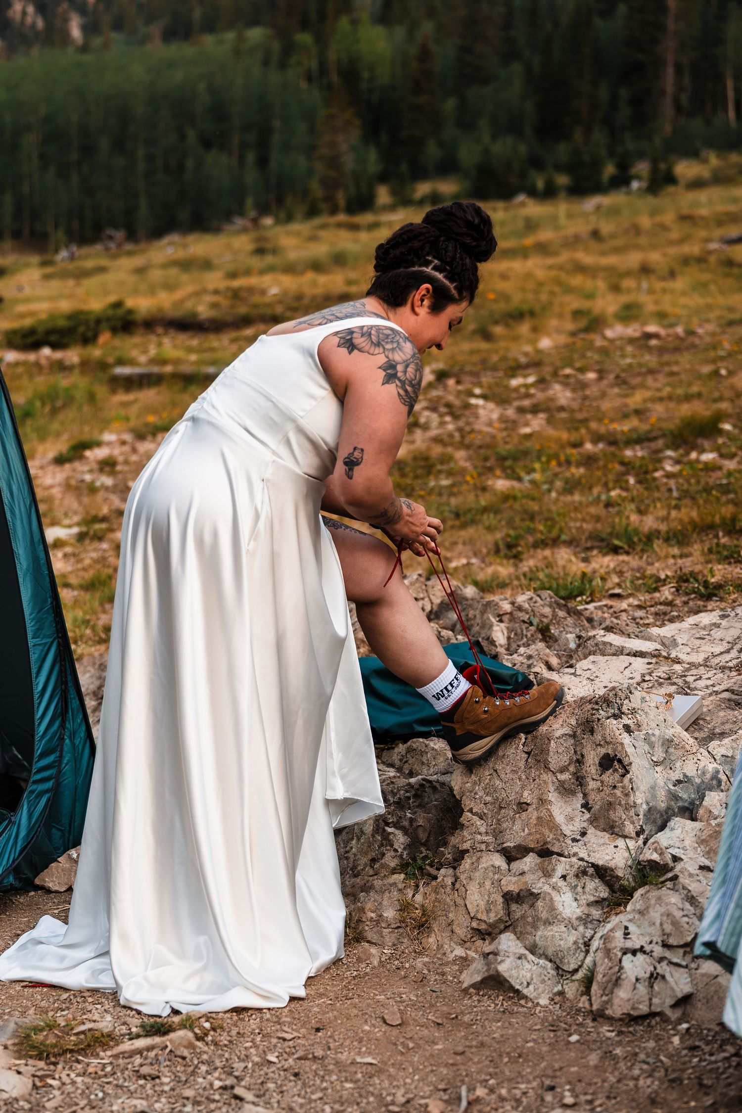 Bride in white wedding gown with arm tattoos adjusts child's shoes on rocky mountain trail during outdoor elopement.