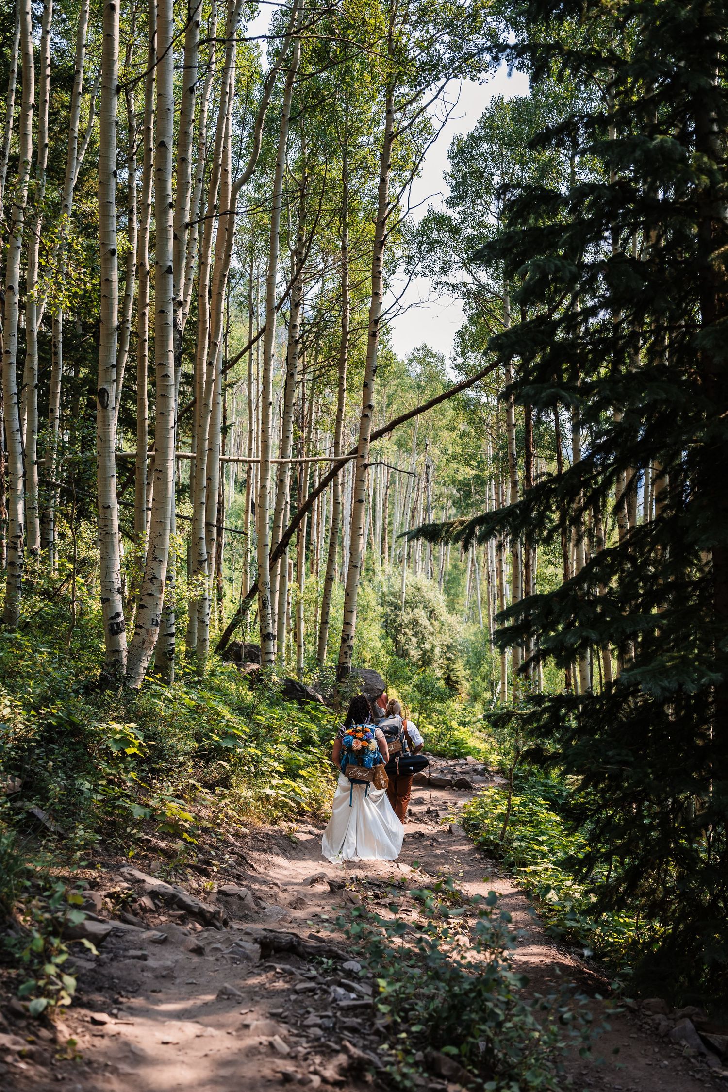 Bride in white wedding dress hikes a rocky forest trail through tall aspen trees with a companion.