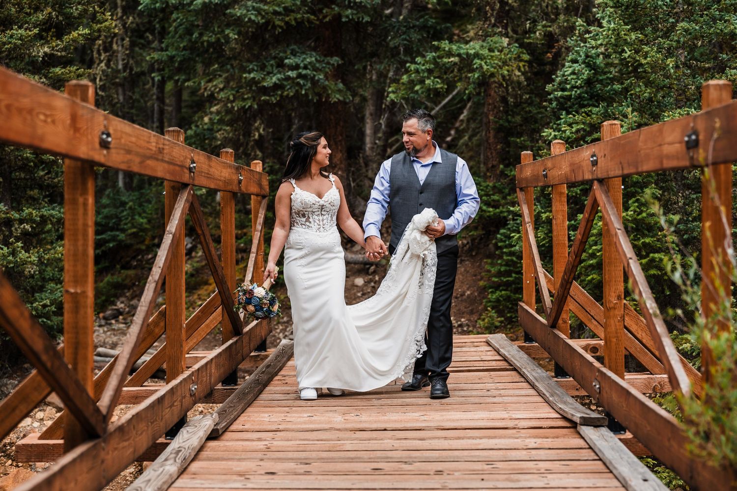 Bride and groom walk hand-in-hand across a rustic wooden bridge surrounded by forest.