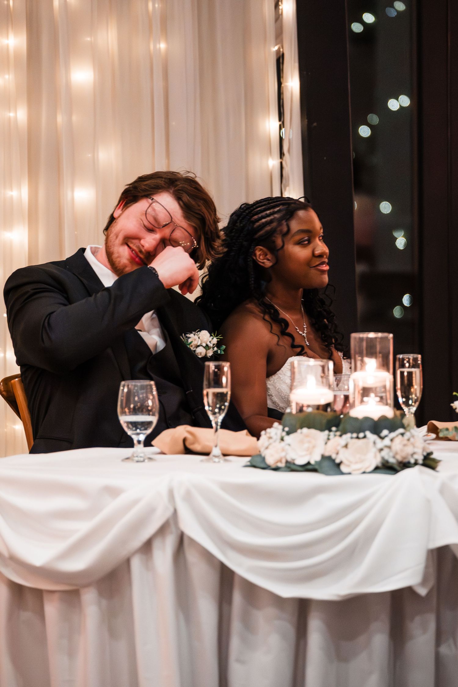 Emotional groom wiping tears beside smiling bride at candlelit wedding reception sweetheart table.
