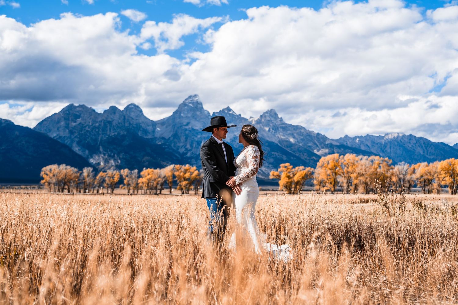 Panoramic wedding photo in golden field with Grand Teton mountains and blue sky in background.