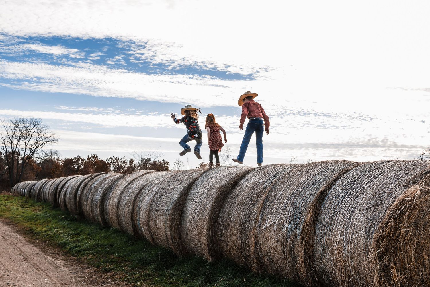 People walking along a line of large round hay bales in a snowy winter field under cloudy skies.