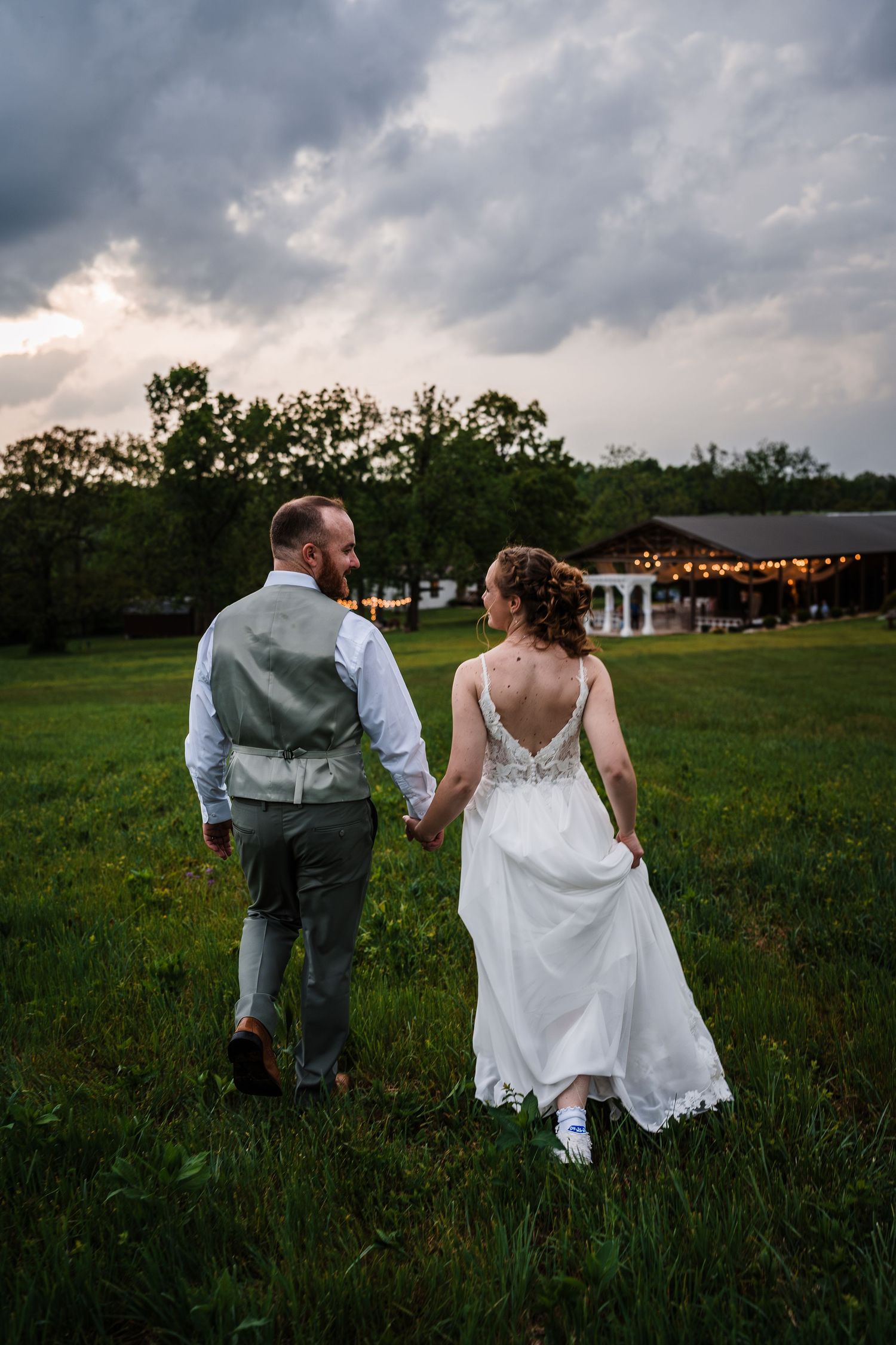 Bride and groom walking hand in hand through green meadow at sunset with wedding venue in background.