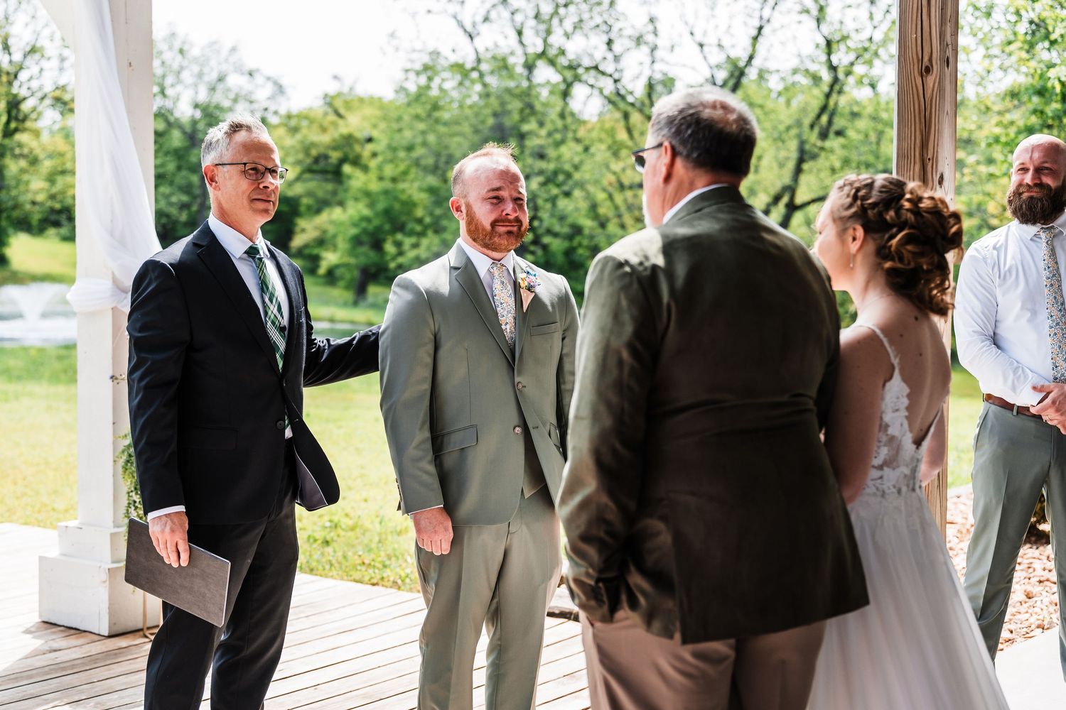 Groom in gray suit smiling with wedding guests on outdoor deck during a sunny wedding ceremony.