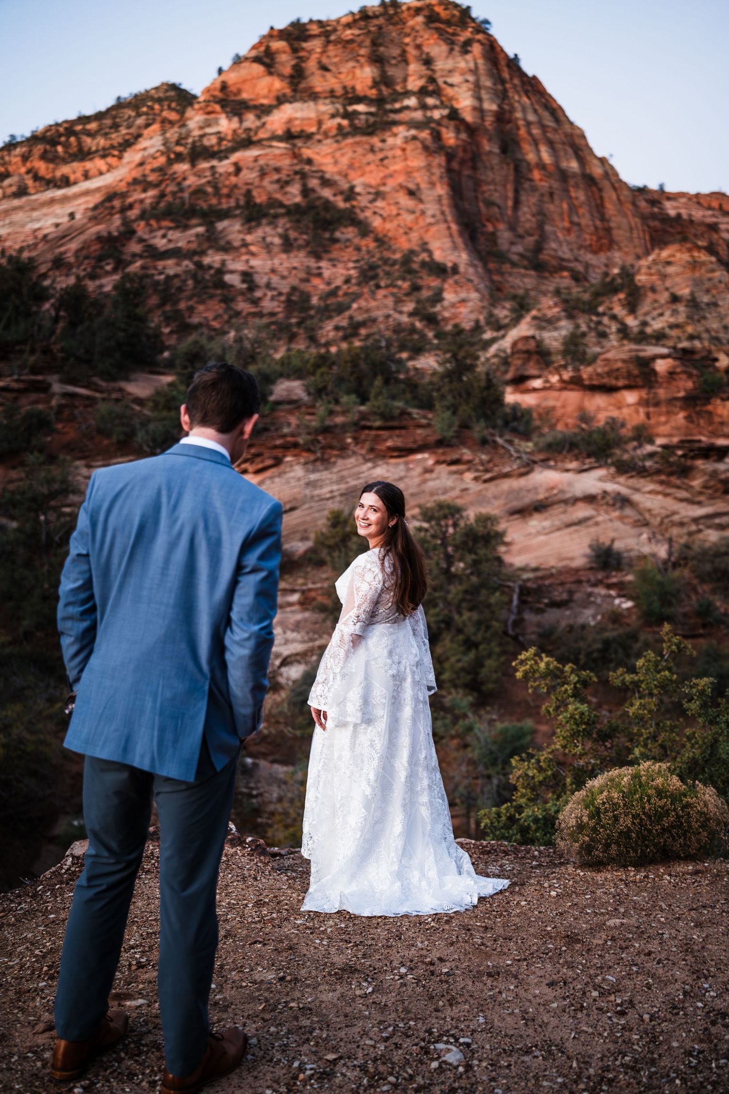 Bride in lace gown looks back at groom in blue suit with red rock canyon backdrop at Zion National Park.
