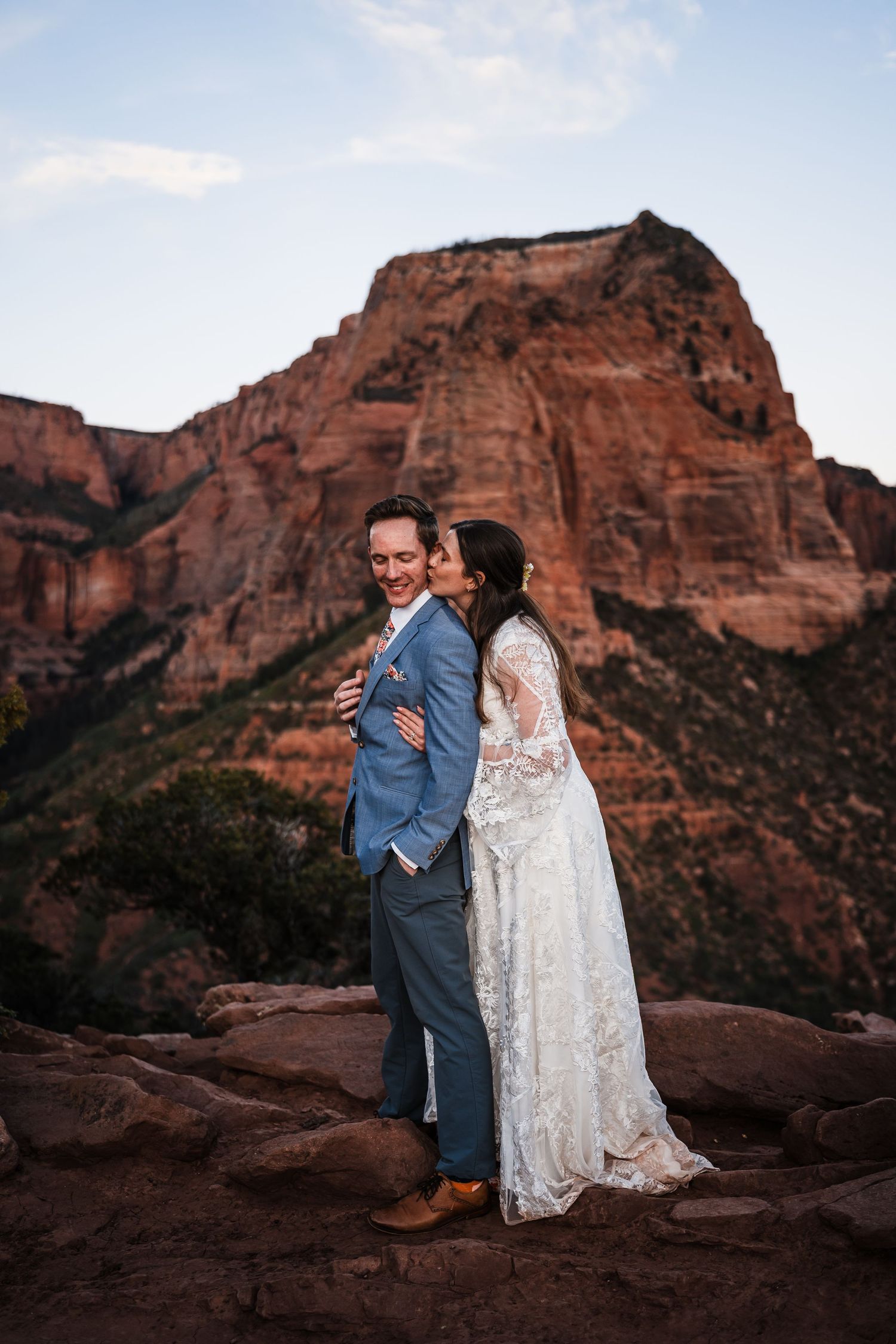 Bride in lace gown kissing groom in blue suit with red rock canyon backdrop at sunset.