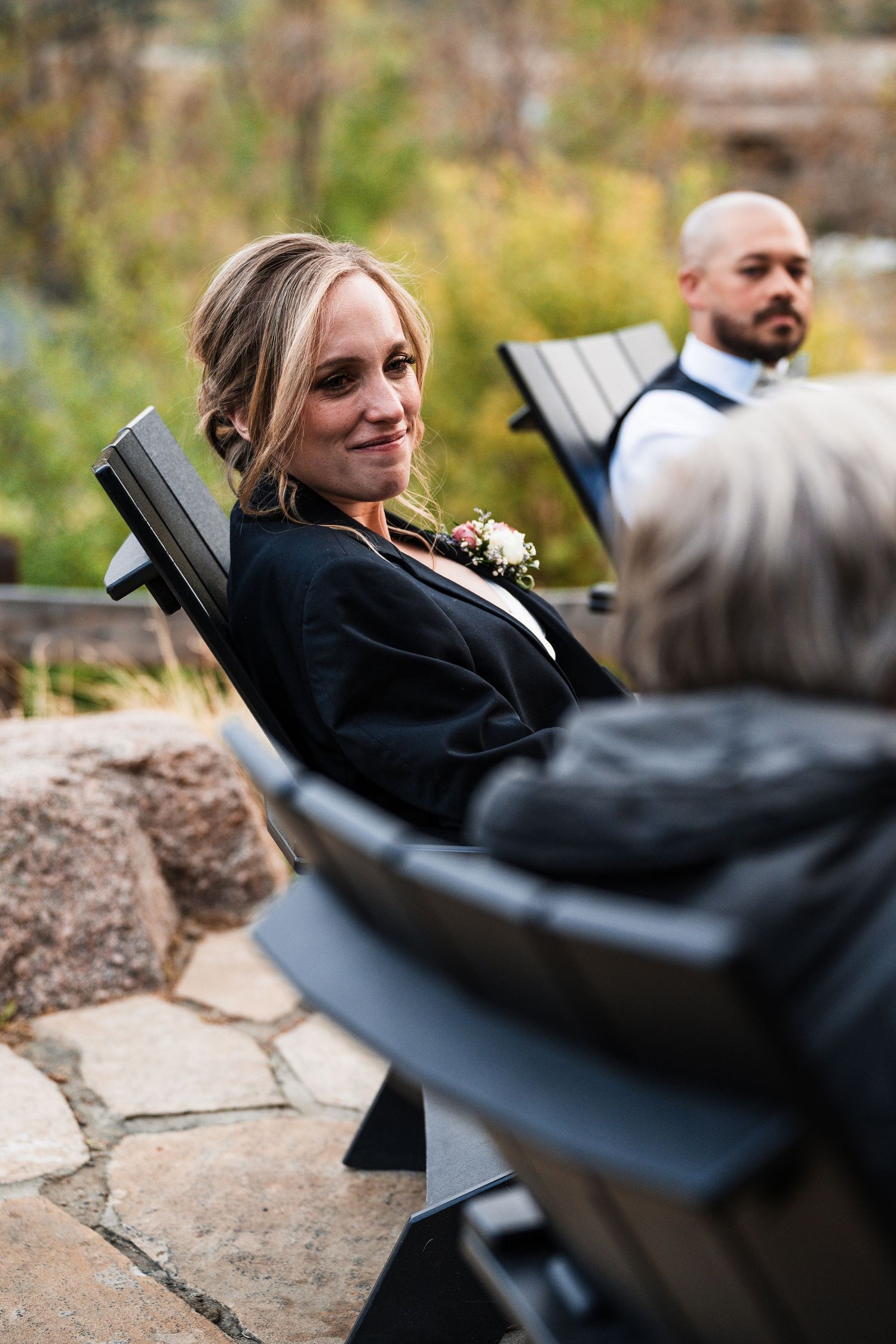 Smiling wedding guest in black jacket with floral corsage seated at outdoor fall wedding ceremony.