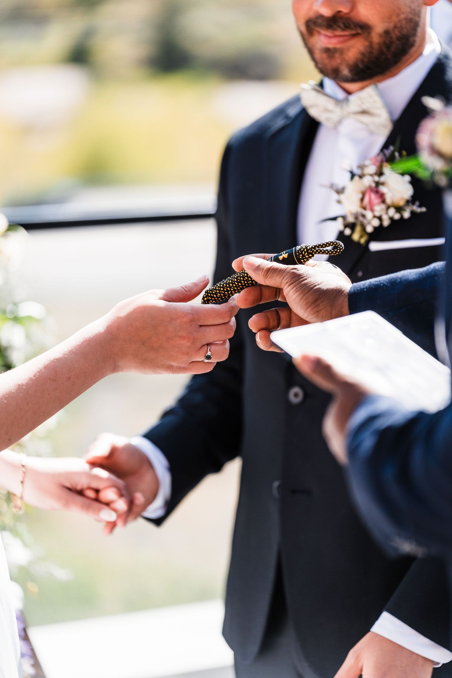 Groom in navy suit with boutonniere receiving wedding rings during outdoor ceremony on sunny day.