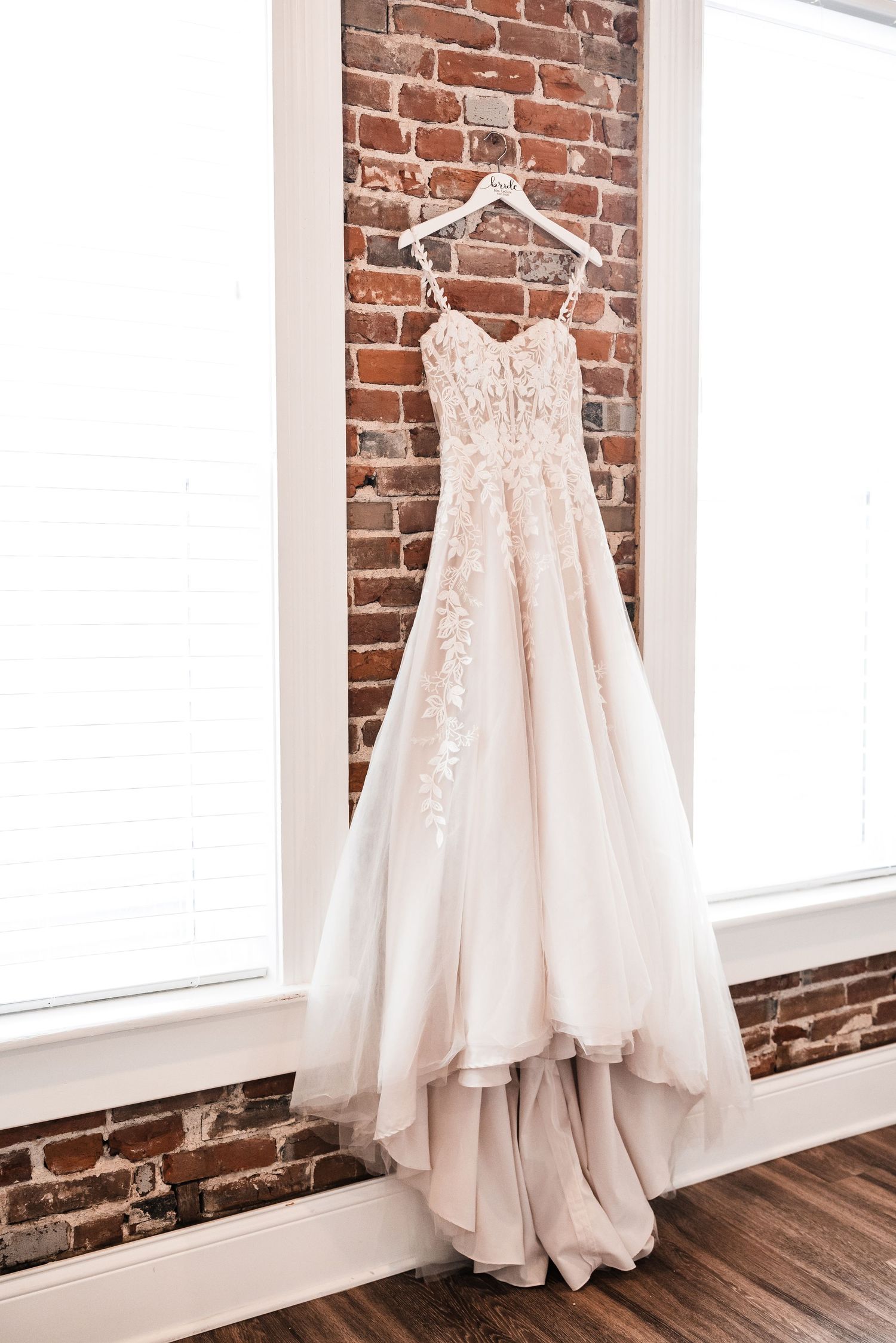 A beautiful white wedding dress with delicate lace details hangs elegantly against an exposed brick wall near a window.