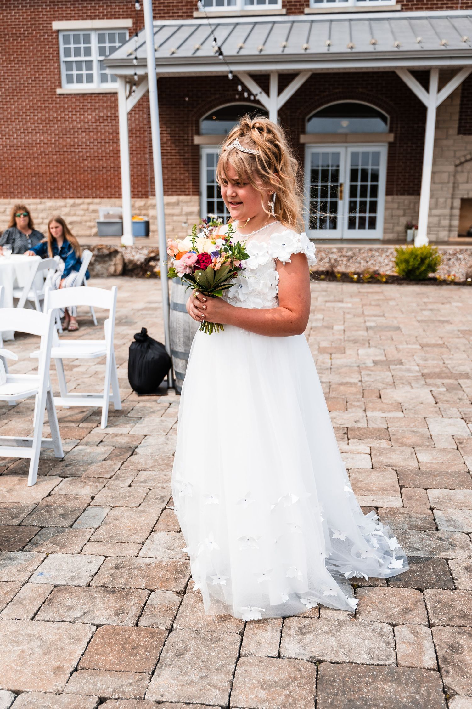 A bride in a flowing white lace gown holds a vibrant bouquet while standing on stone pavers before a brick building.