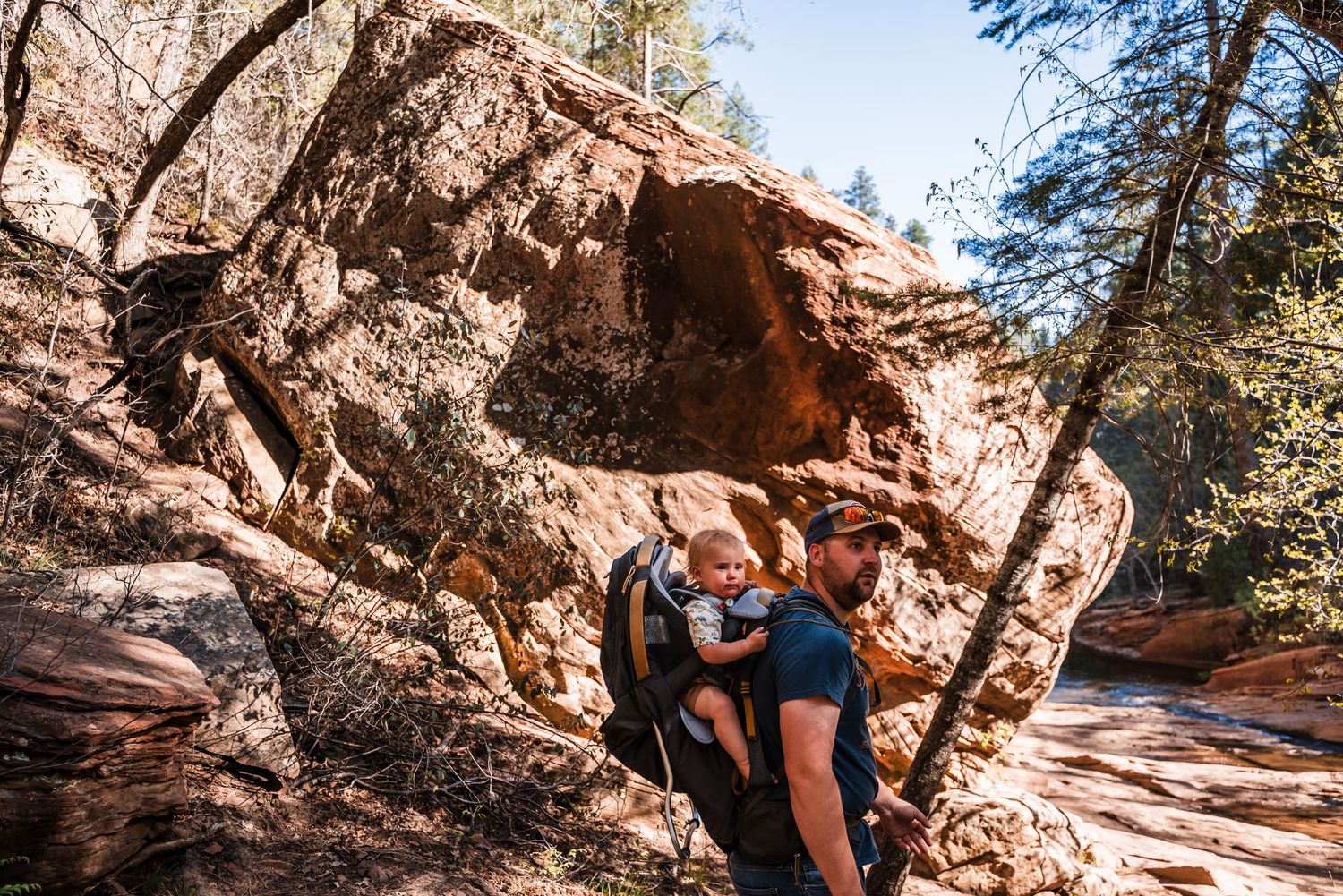Hiker resting beneath large red rock formations and overhanging cliffs on a desert trail.