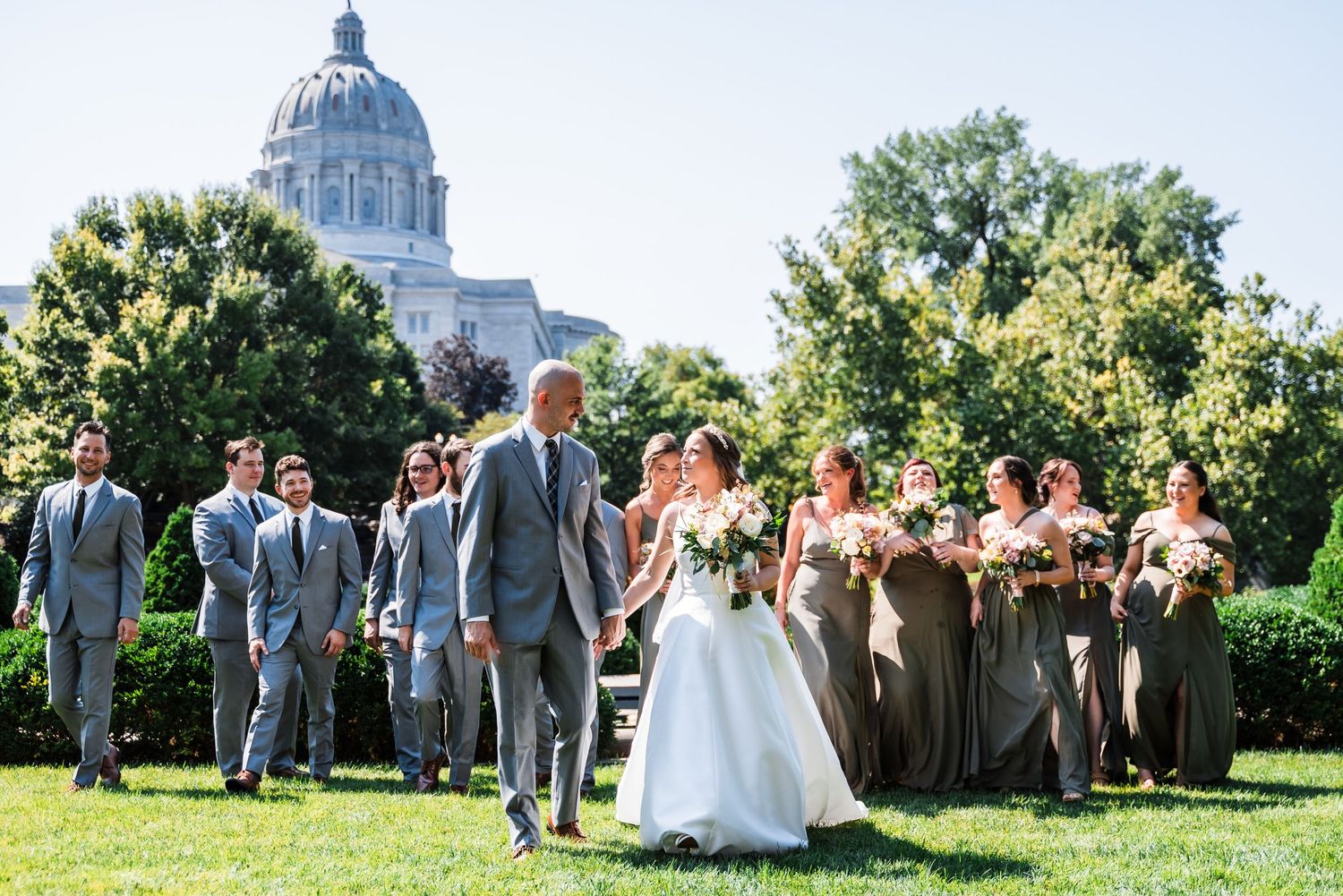 Wedding party poses in front of state capitol building on sunny summer day with green trees.