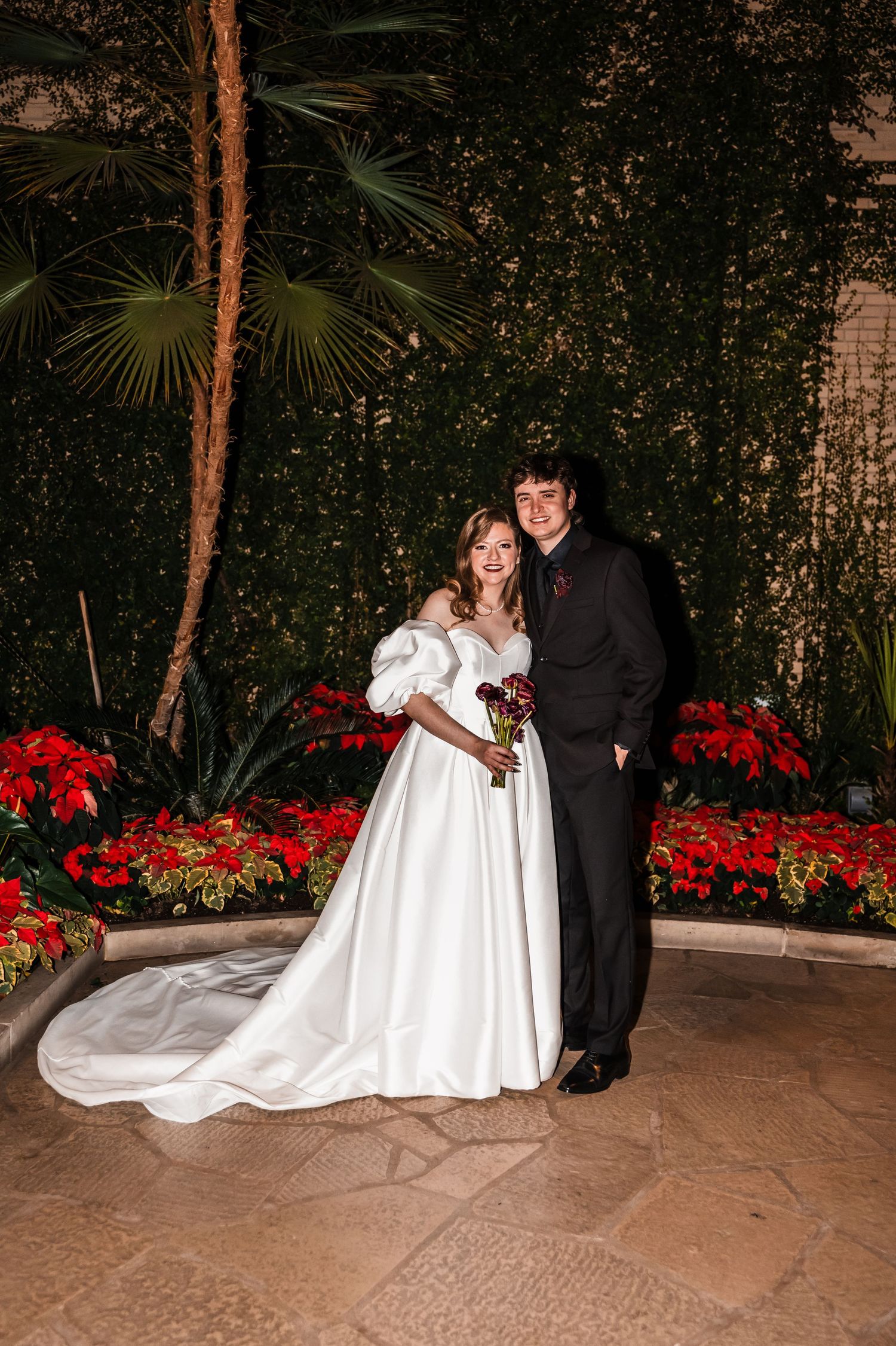 Couple poses for wedding portrait in front of red poinsettias and tropical foliage.