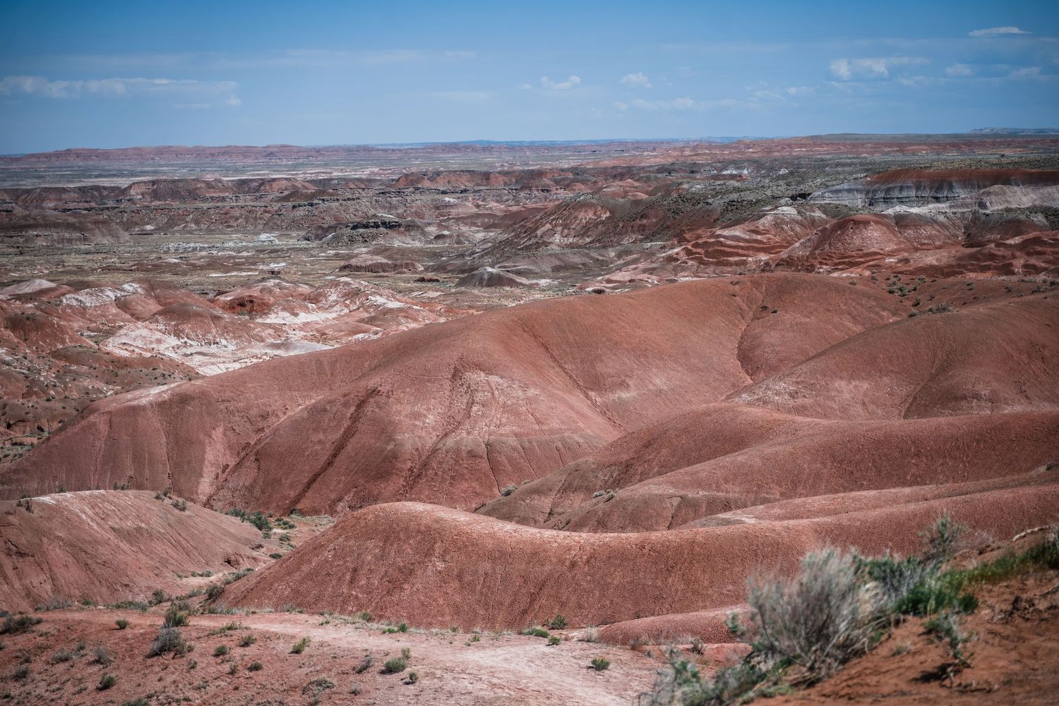 Red and orange layered badlands stretch across an arid desert landscape beneath a bright blue sky.