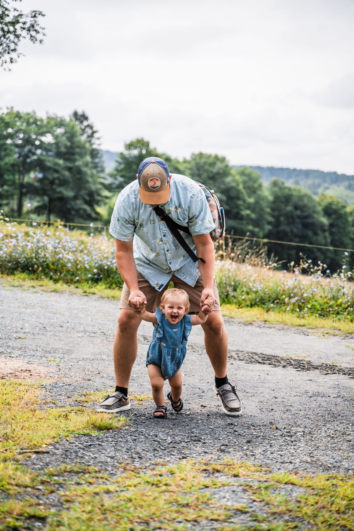 Joyful playtime on a gravel path with a small child in overalls being held up against mountain scenery.