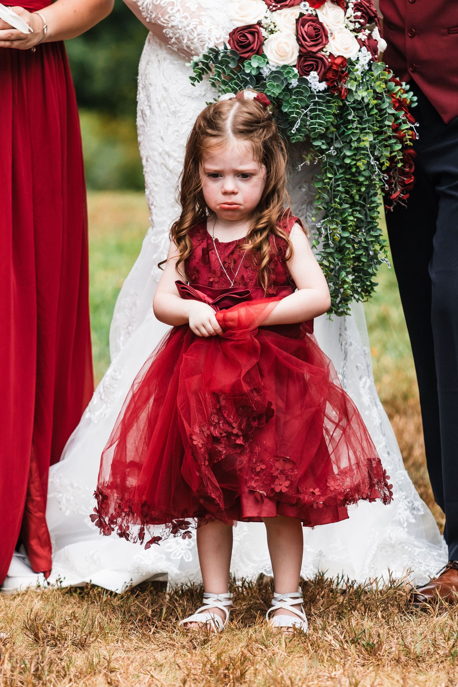 A flower girl in a red tulle dress stands at a wedding ceremony with white and burgundy decor.