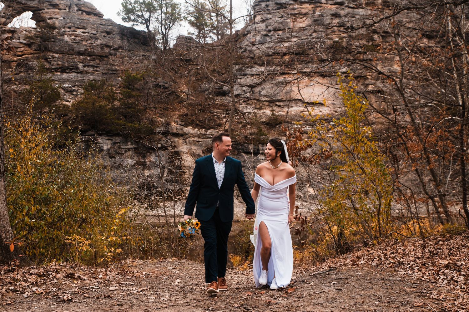 A couple walks hand in hand on a path with towering rock formations and autumn colors.