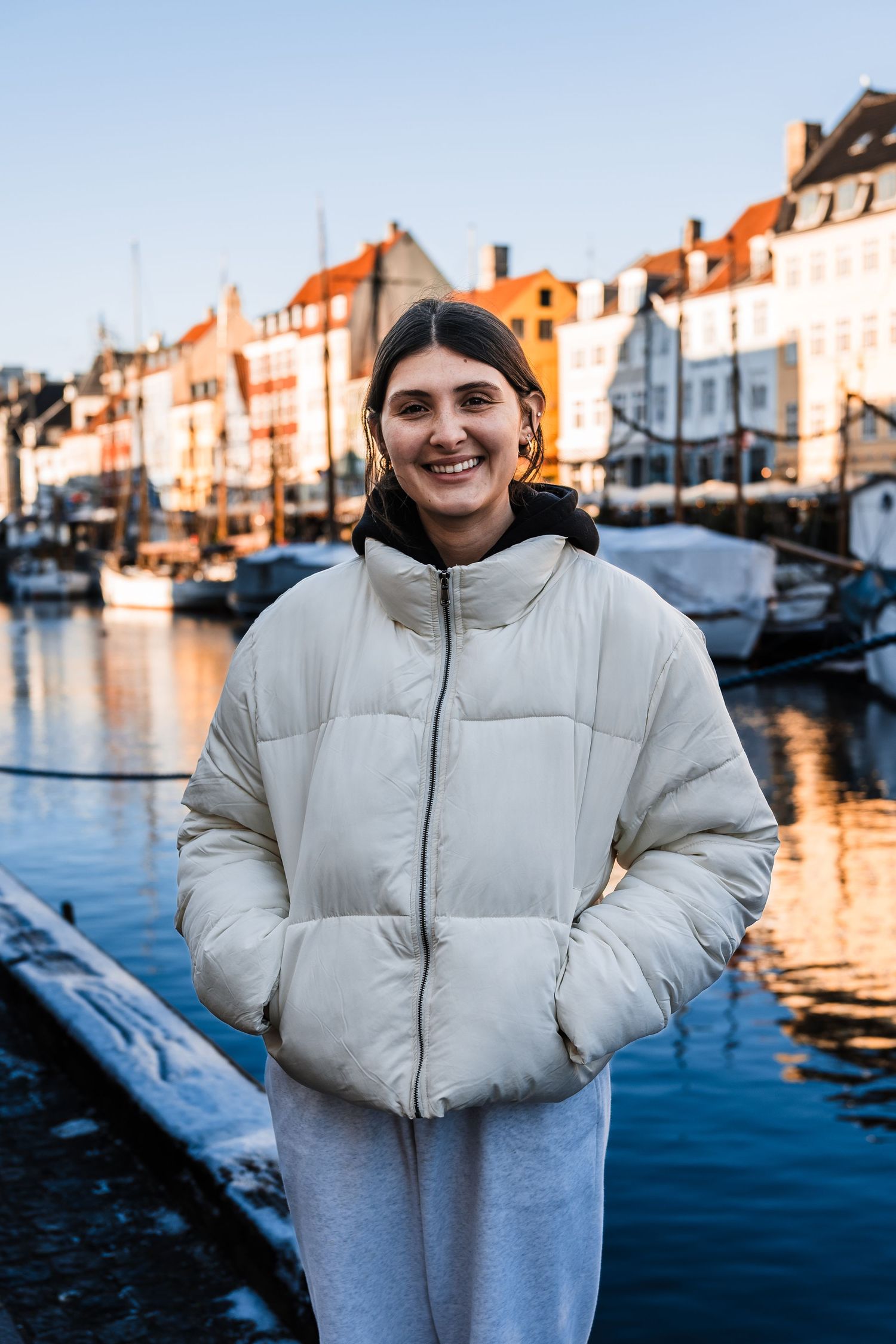 Person in white puffer jacket stands along scenic waterfront with boats and buildings at sunset.