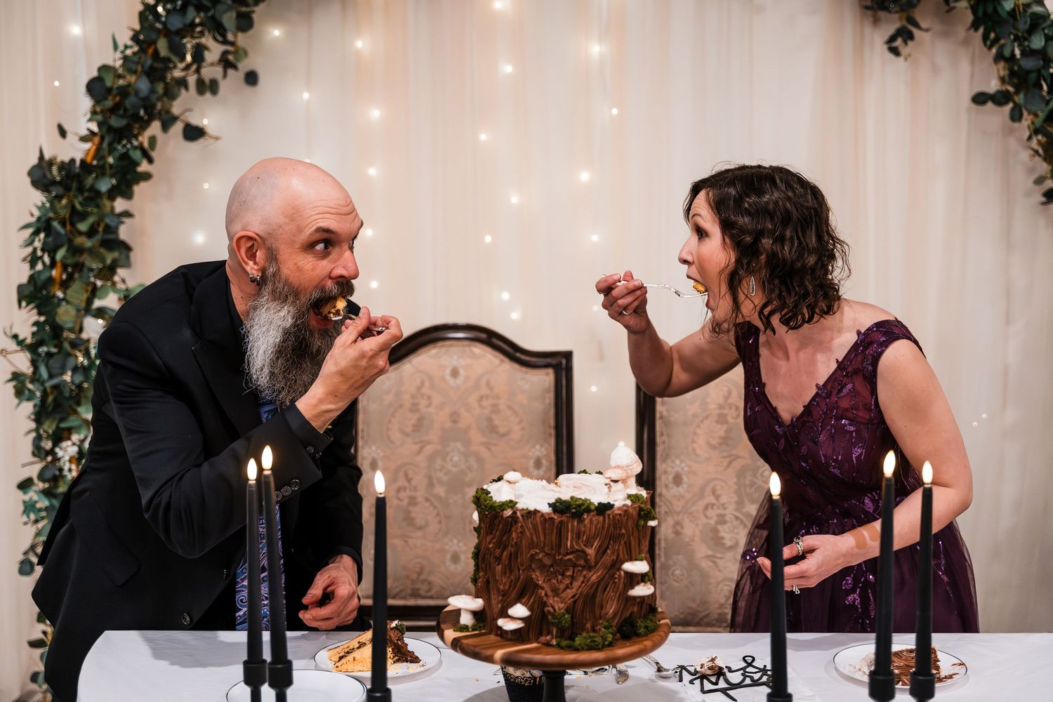 Bride and groom playfully feeding each other rustic woodland wedding cake at reception table.