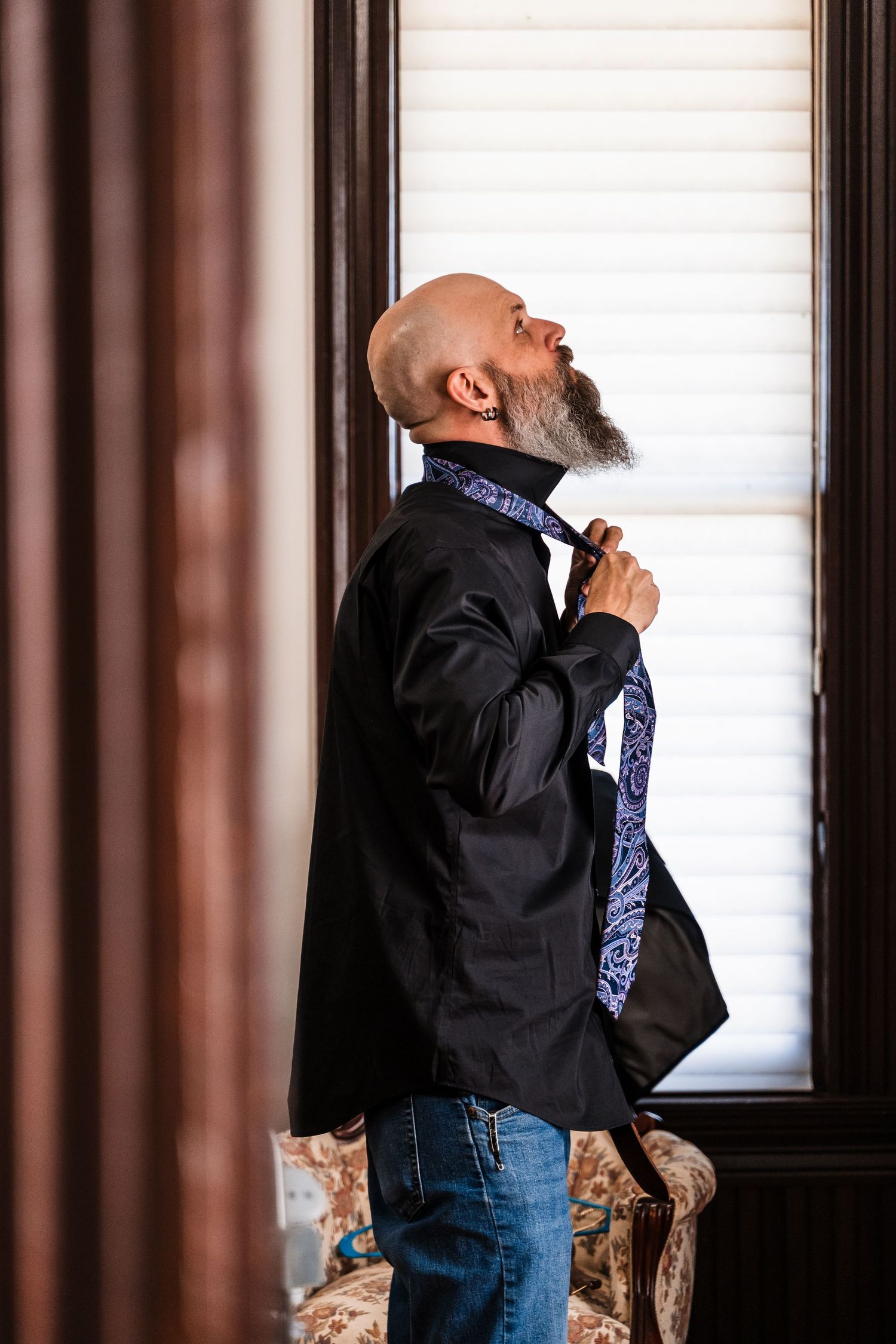 Bald man with gray beard adjusting blue paisley tie wearing black blazer and jeans standing by window.