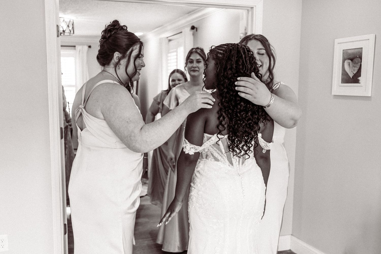 Black and white photo of bridesmaids helping bride into wedding dress in a bright room.