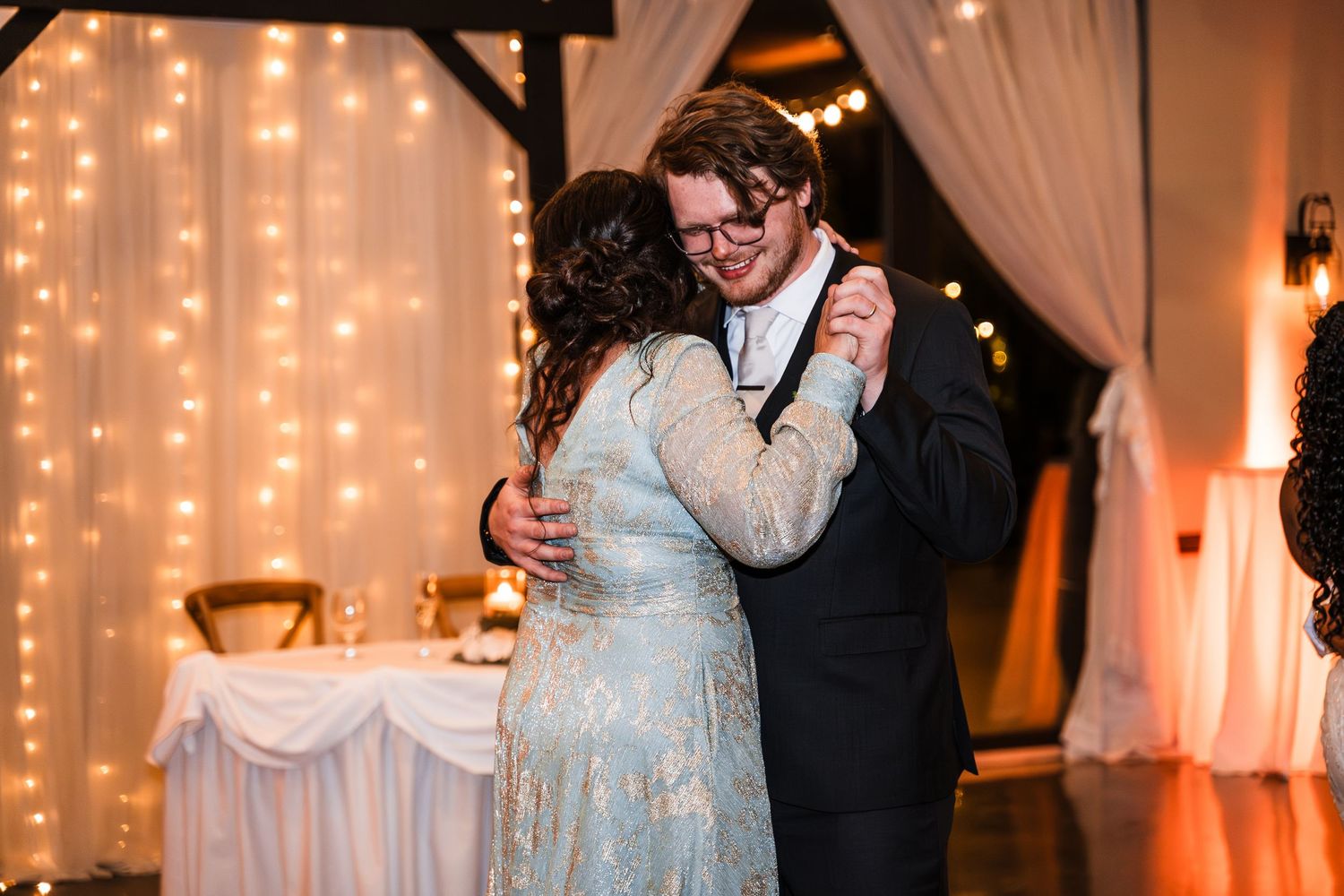 Groom dancing with woman in blue floral gown under warm string lights at wedding reception.