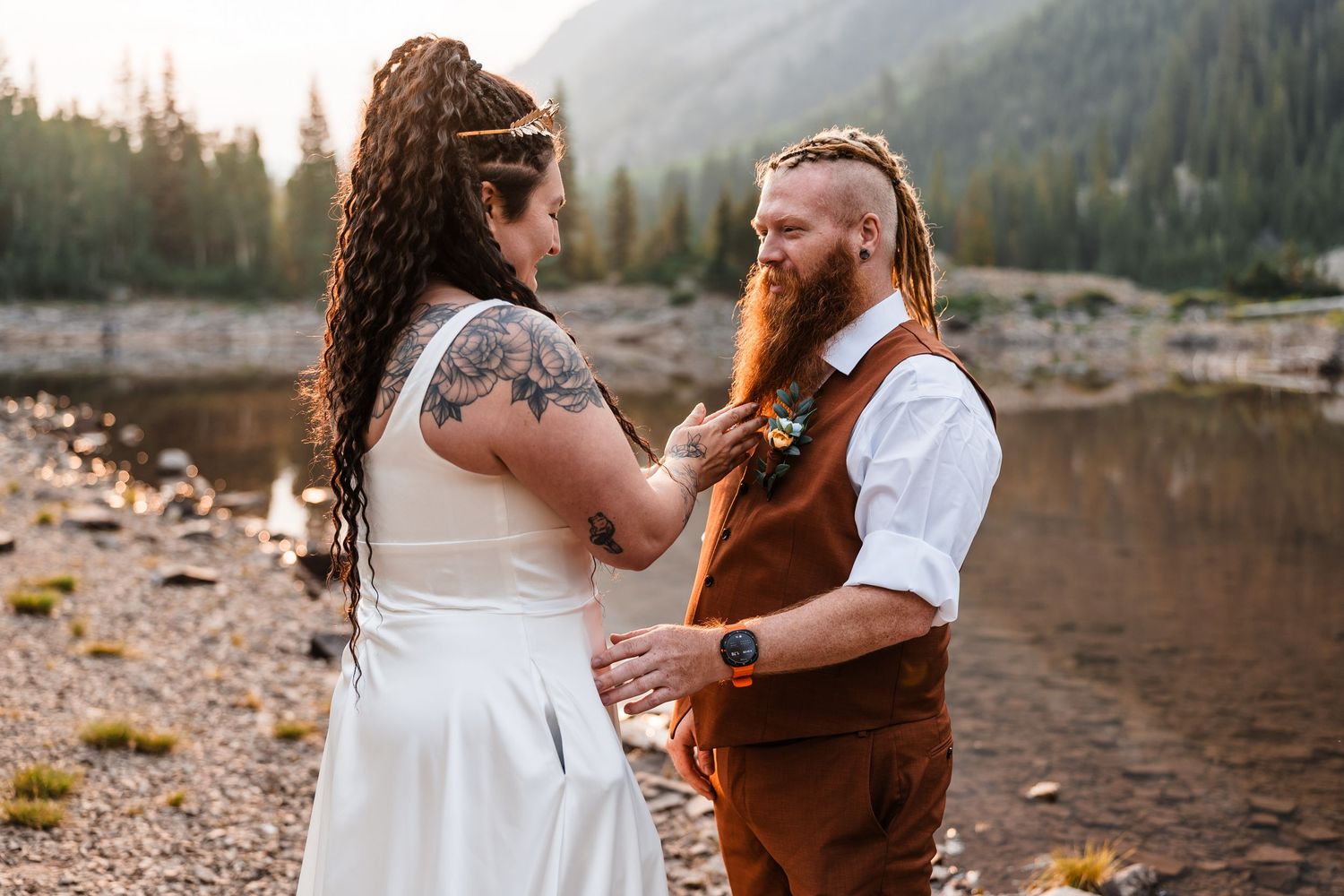 Bride with tattoos adjusting groom's boutonniere by mountain lake during outdoor wedding ceremony.