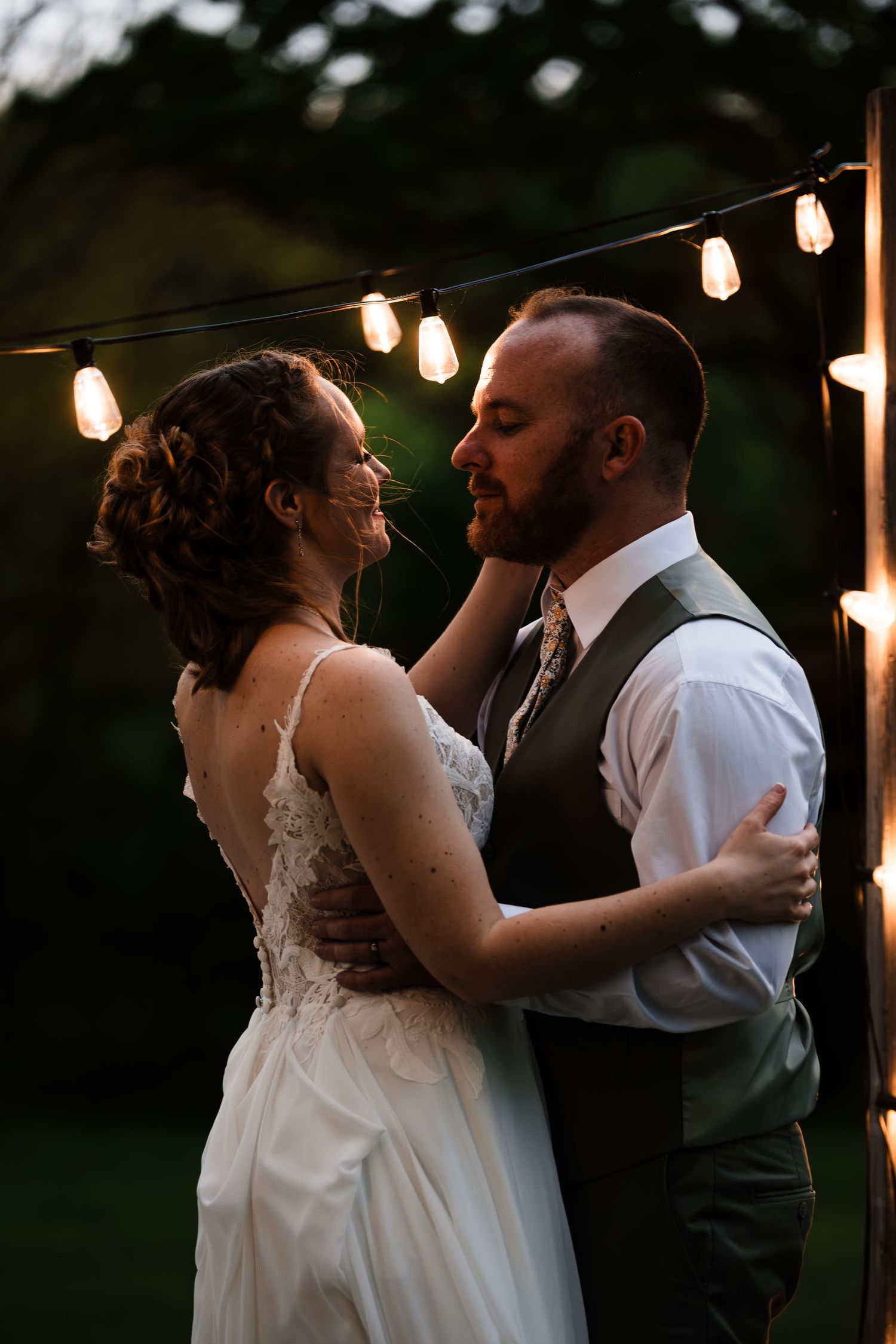 Bride and groom share an intimate moment under warm string lights at dusk in outdoor wedding setting.