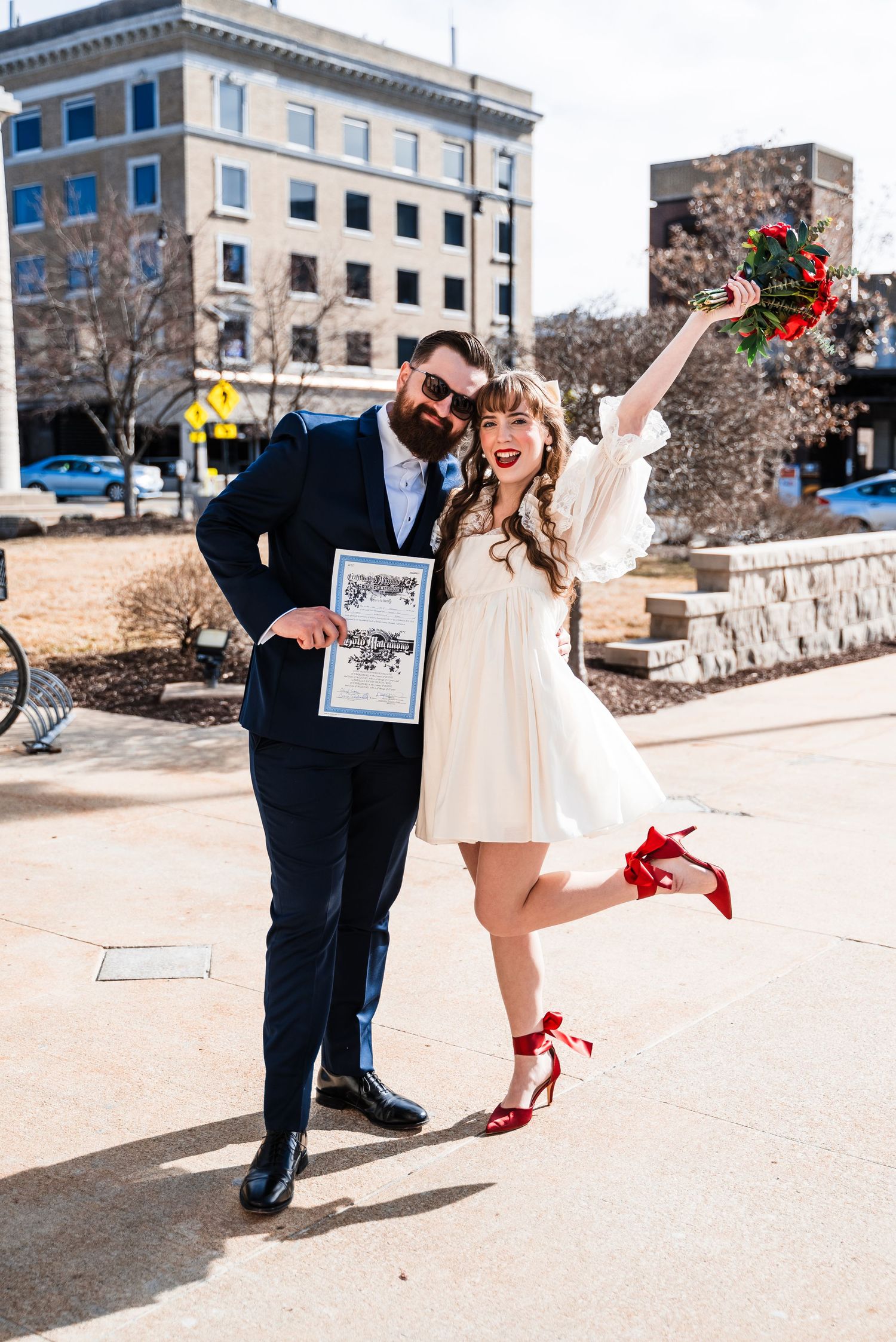 A couple celebrates their graduation with playful poses on a sunny day outside a university building.