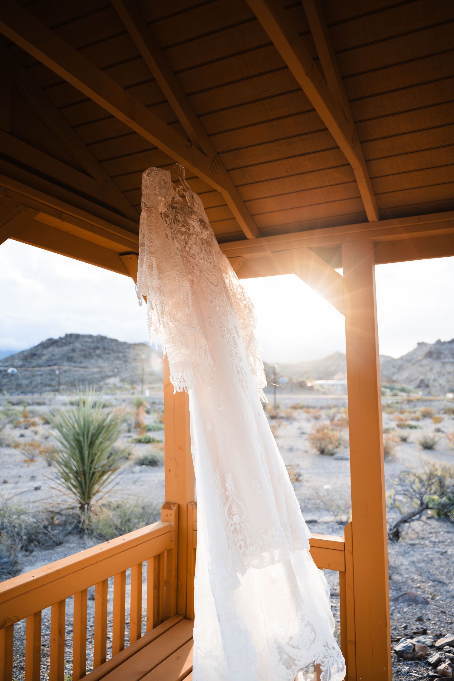 A delicate white lace wedding dress hangs on a wooden porch with desert mountains and sunset in the background.