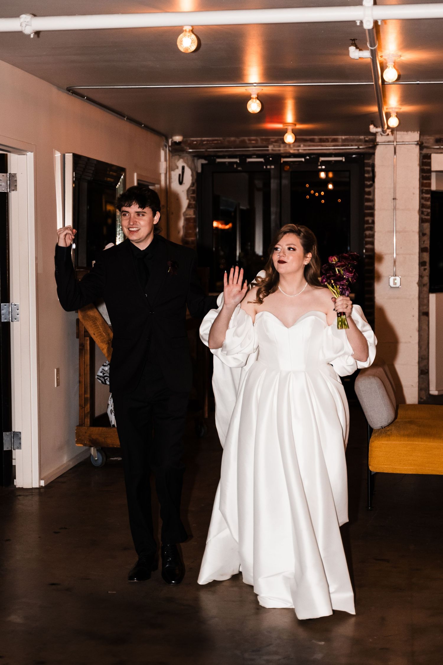 Couple gestures enthusiastically while walking through indoor venue with string lights overhead.