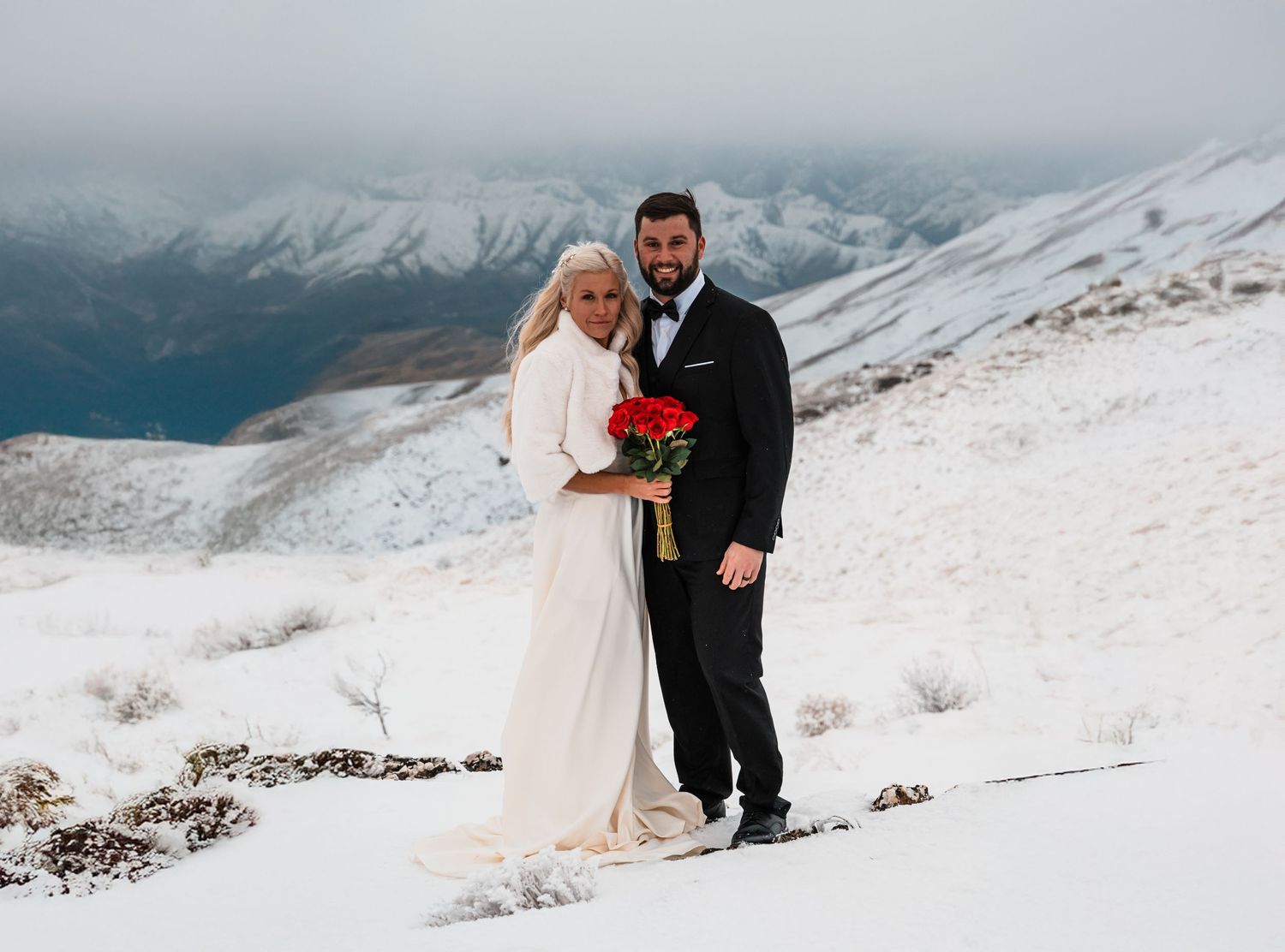 A winter wedding couple embraces on a snowy mountain slope with dramatic snow-capped peaks in the background.