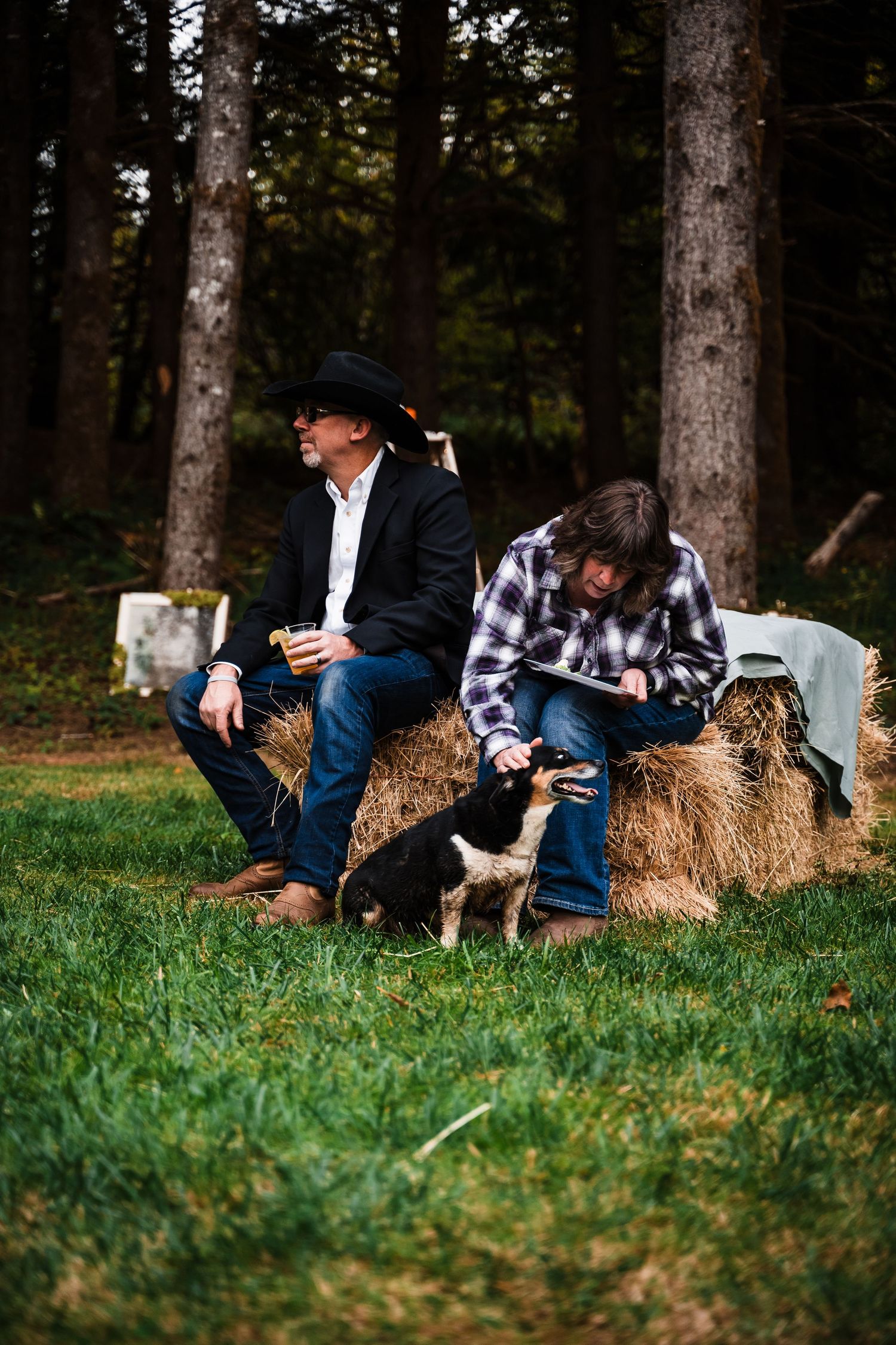 Two people relaxing on hay bales with a small dog in a wooded setting.