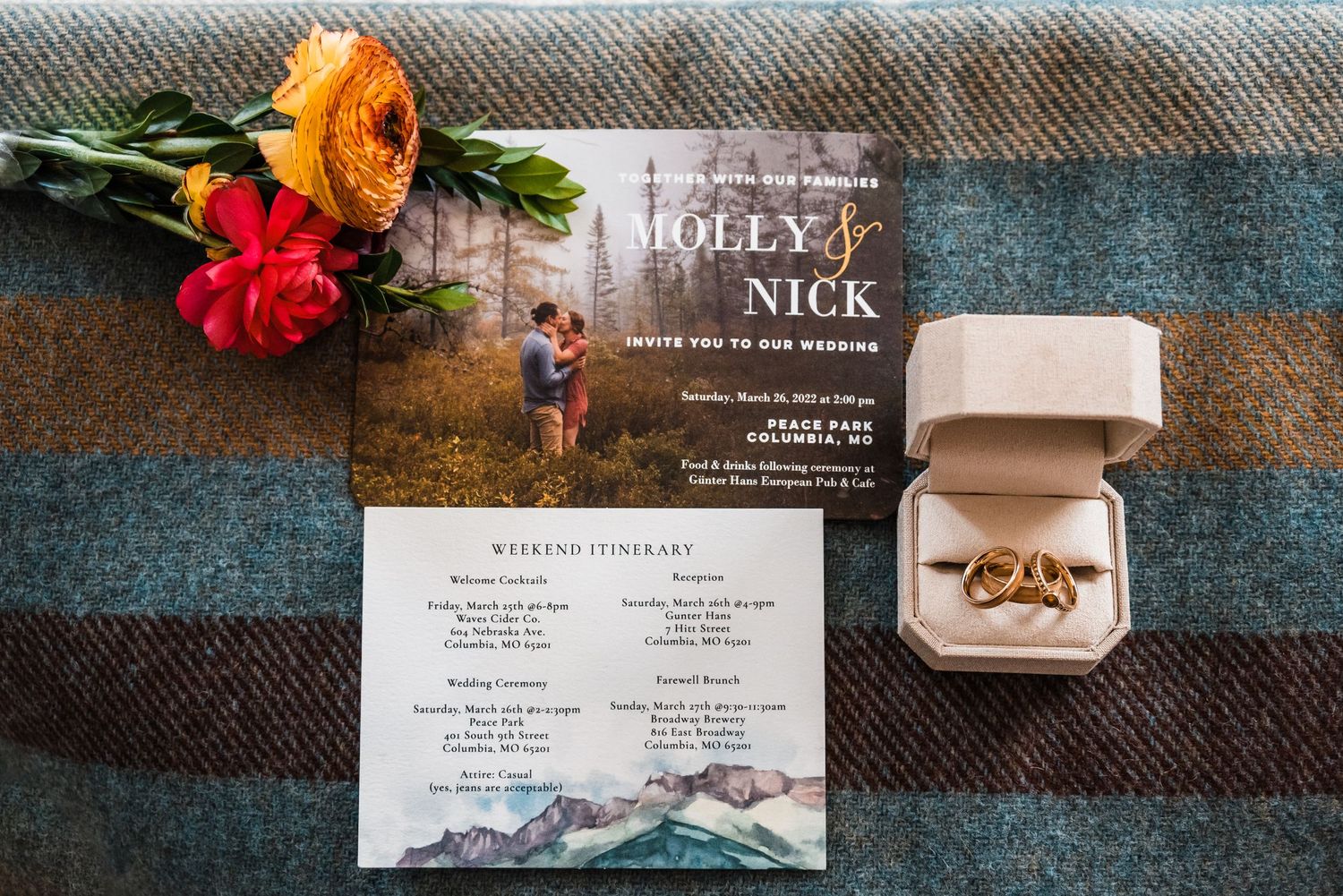 Wedding invitation with orange and red flowers and a ring box on a plaid blanket.