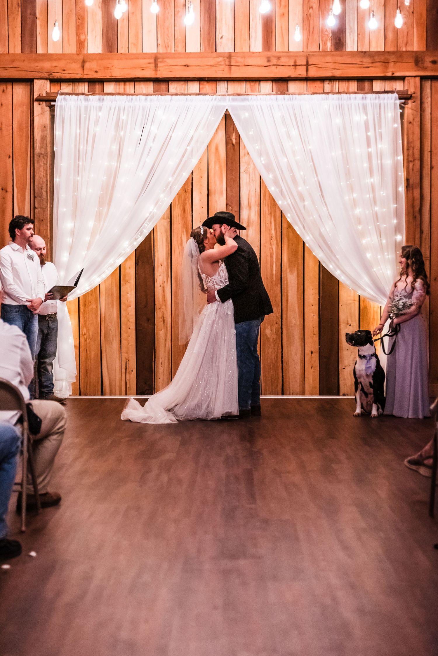A romantic wedding ceremony takes place in a rustic barn setting with white drapes and wooden backdrop.