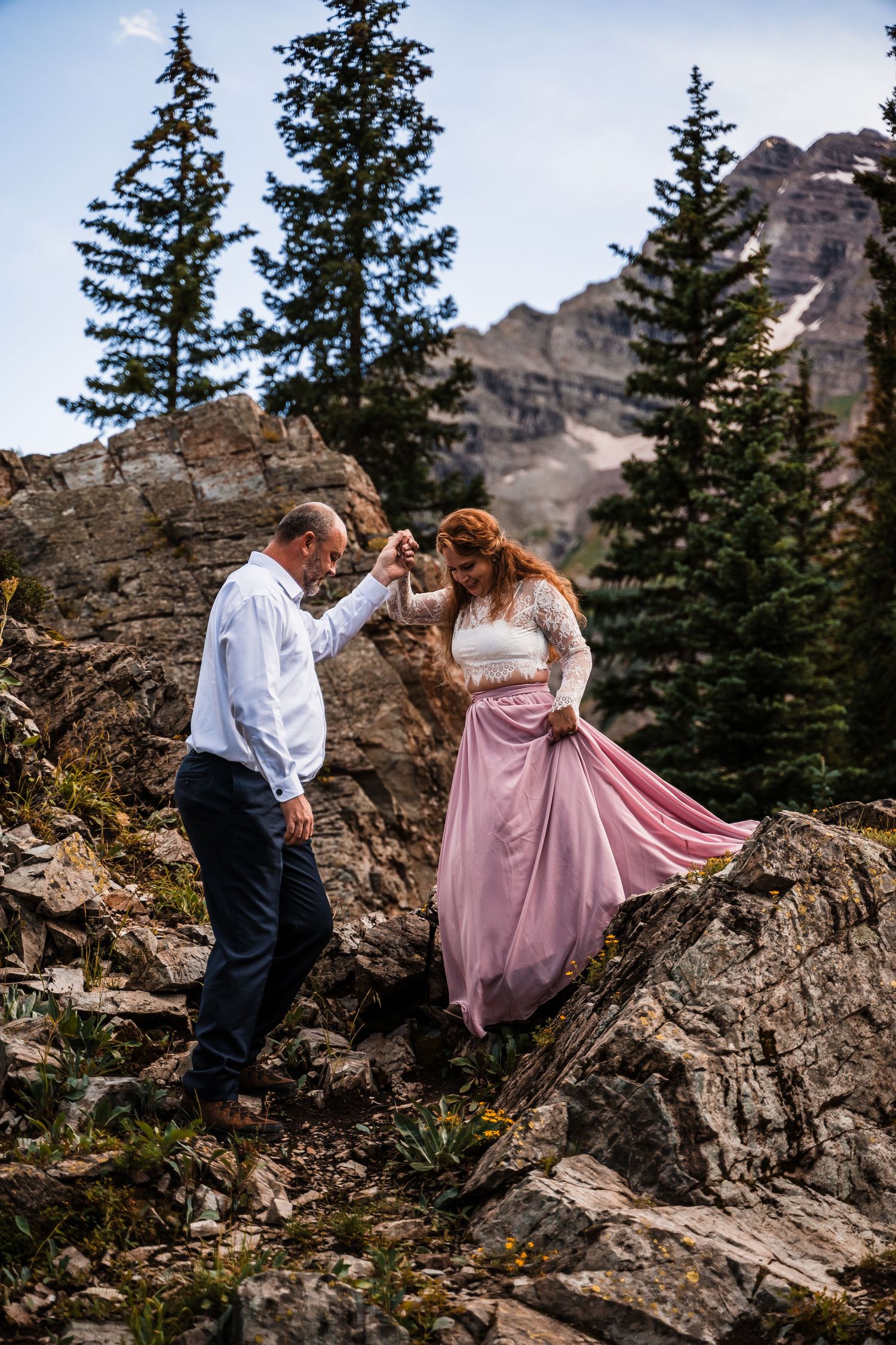 A couple shares a romantic moment on rocky mountain terrain with evergreen trees and peaks in the background.